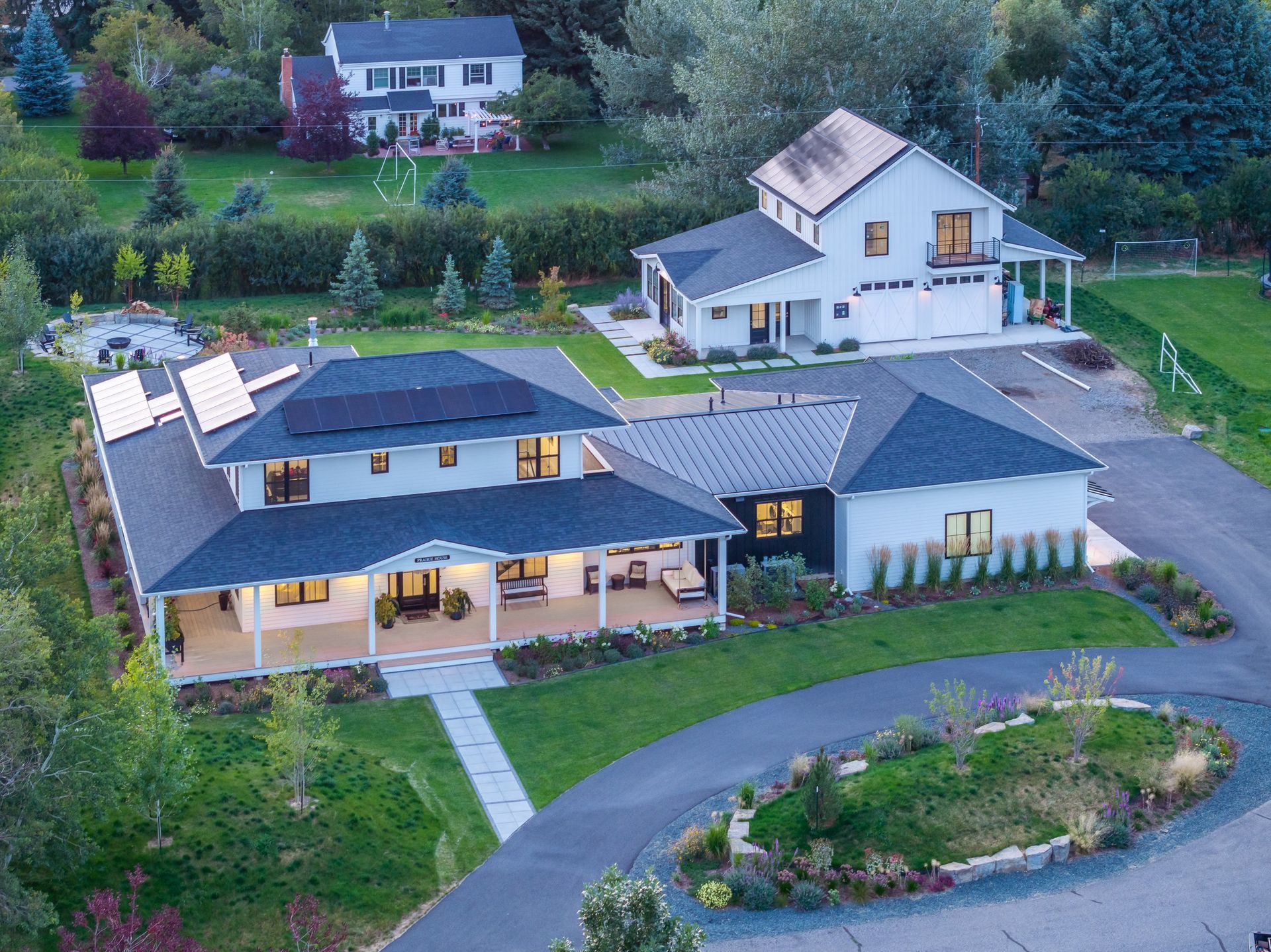 An aerial view of a white, modern farmhouse with a dark roof, solar panels, and a large porch, set in a lush green yard.