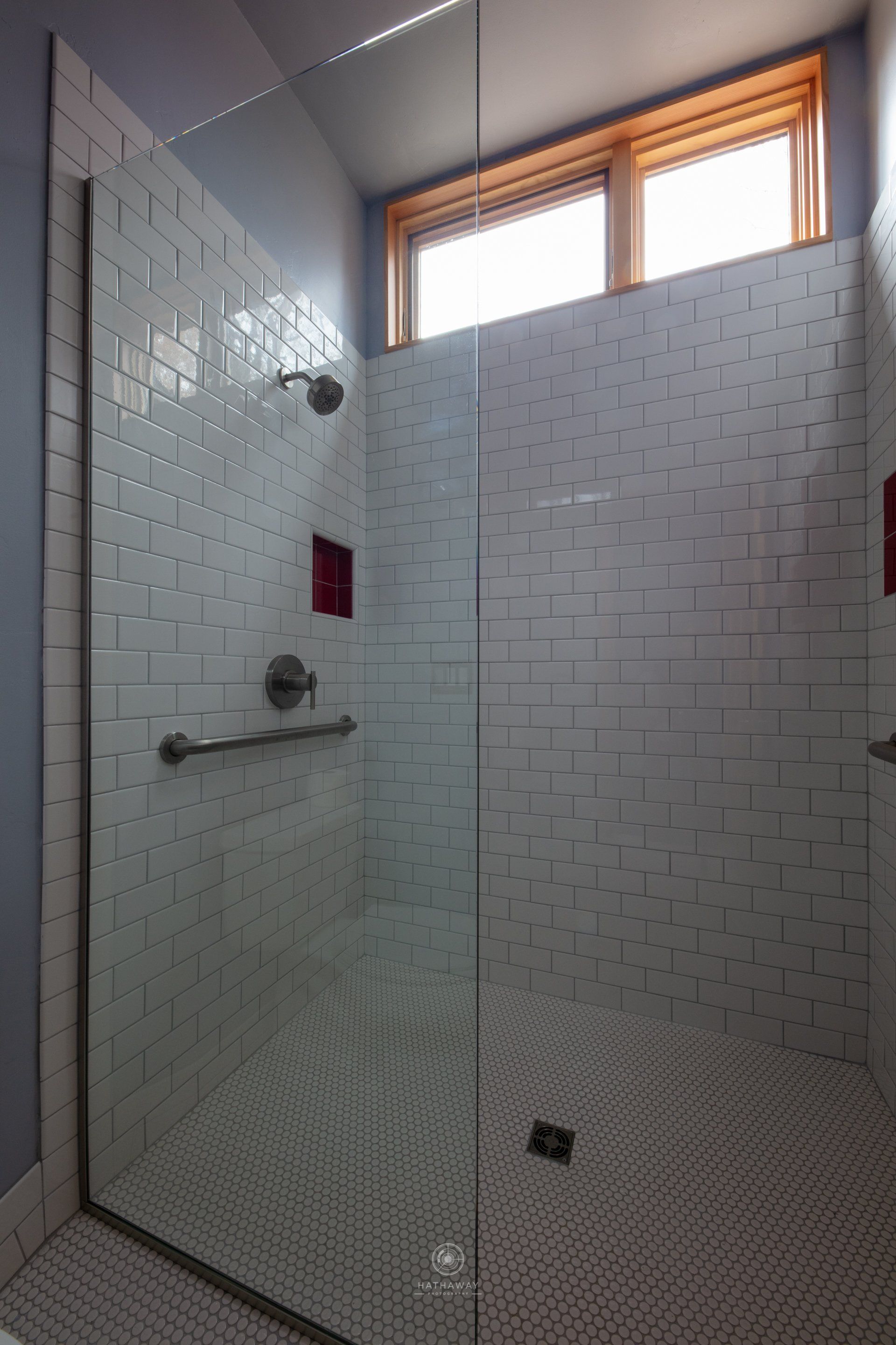 A modern shower with white subway tile walls, a small high window, a glass door, and a pebble-tiled floor.