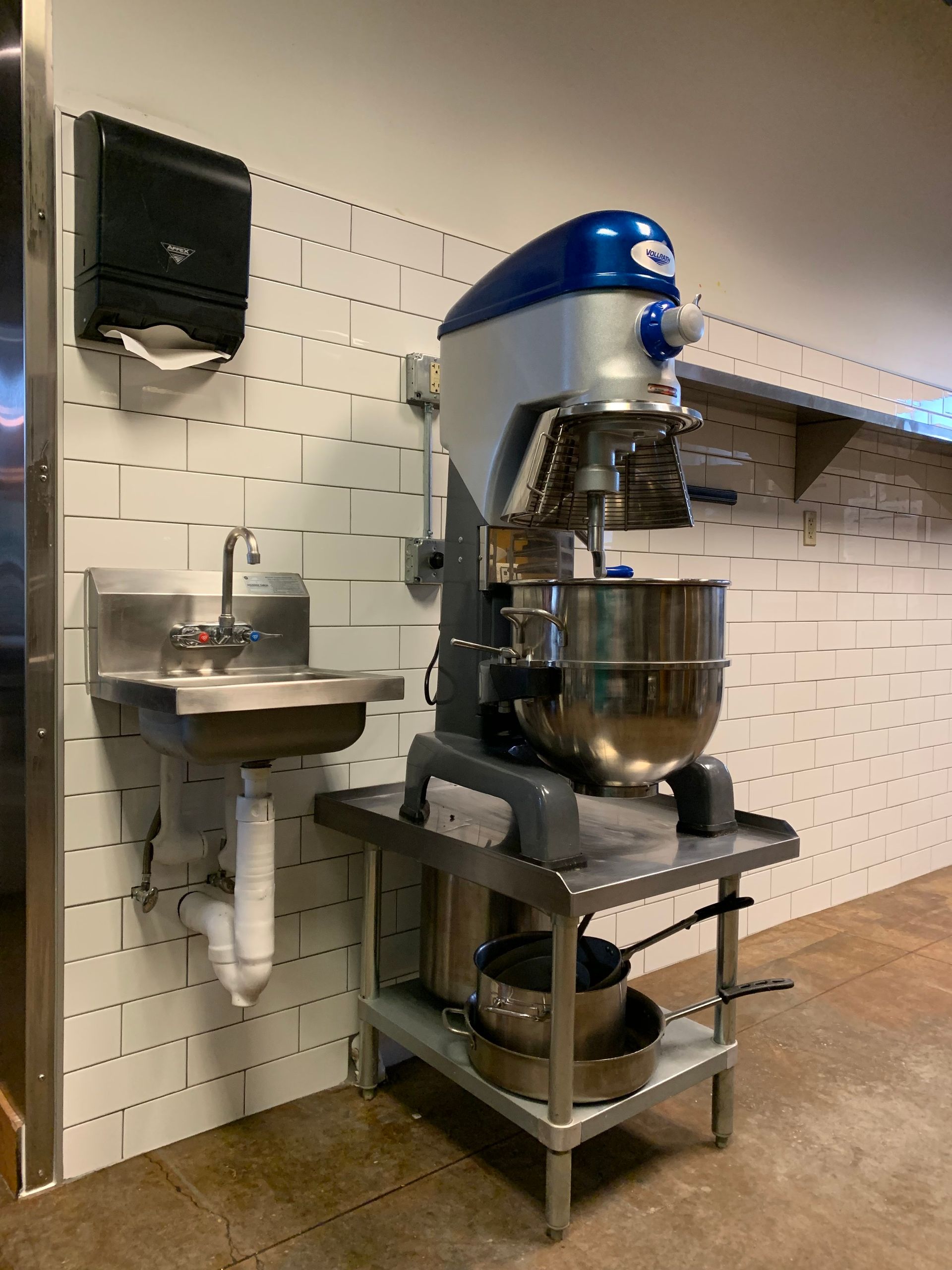 A commercial kitchen setting with a metal sink, a paper towel dispenser, and a large blue and silver standing mixer.