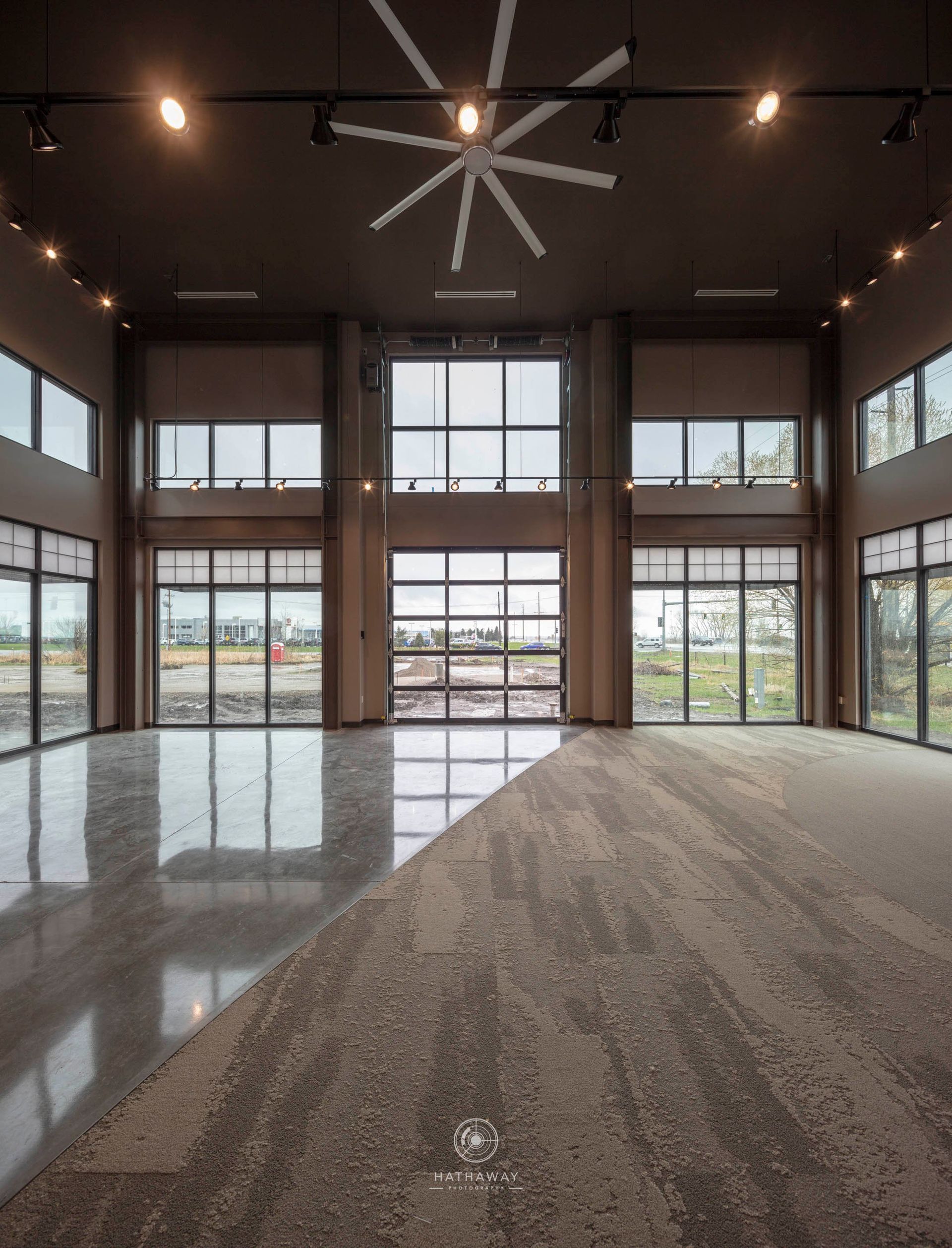 A high-ceilinged room with large windows, divided diagonally by polished concrete and textured carpet flooring.