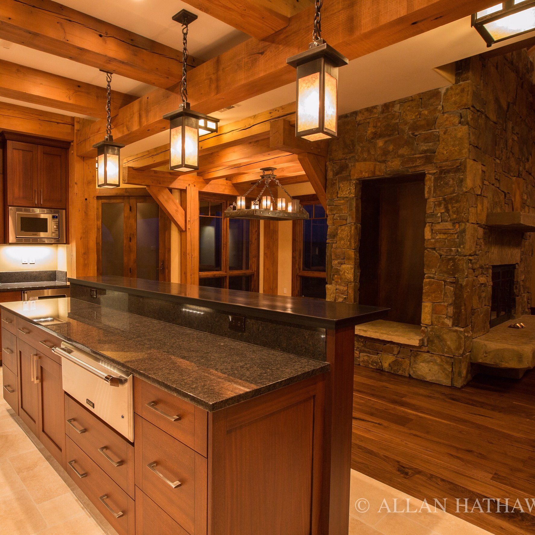 A rustic kitchen with a large wooden island, stone fireplace, exposed timber ceiling beams, and warm pendant lighting.