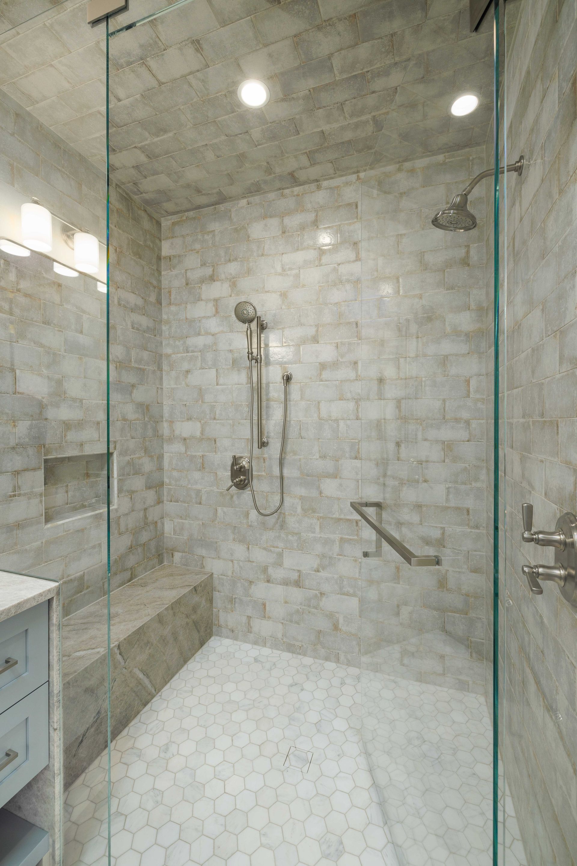 A modern shower with light-toned stone tile walls, a built-in bench, a glass door, and a white pebble floor.