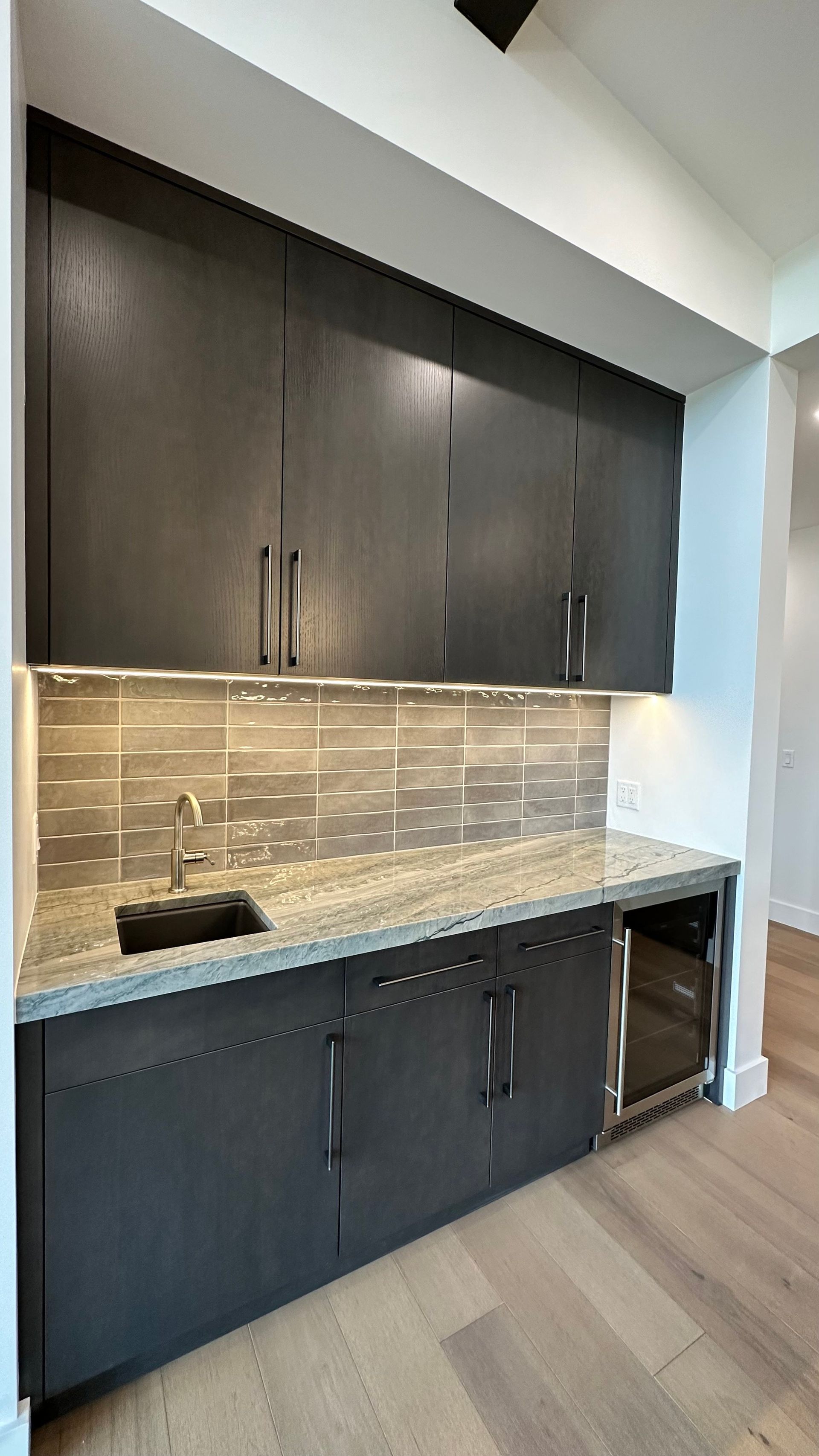 A modern wet bar with dark wooden cabinets, granite countertops, a stainless sink, and a built-in wine cooler.