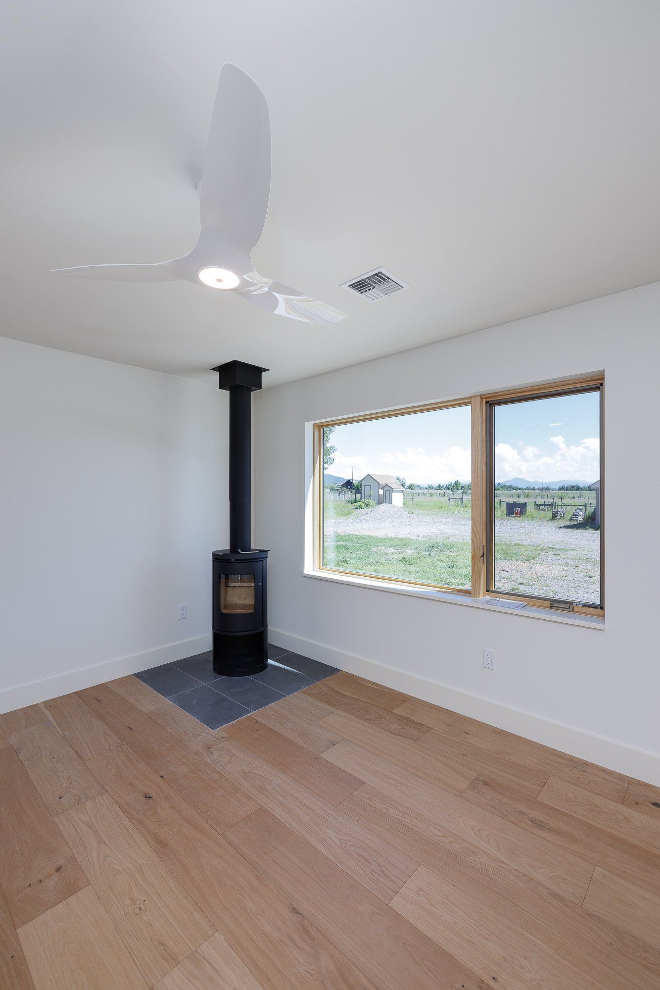 A modern room with white walls, light wood floors, a black wood-burning stove, and a view of an open field through a window.