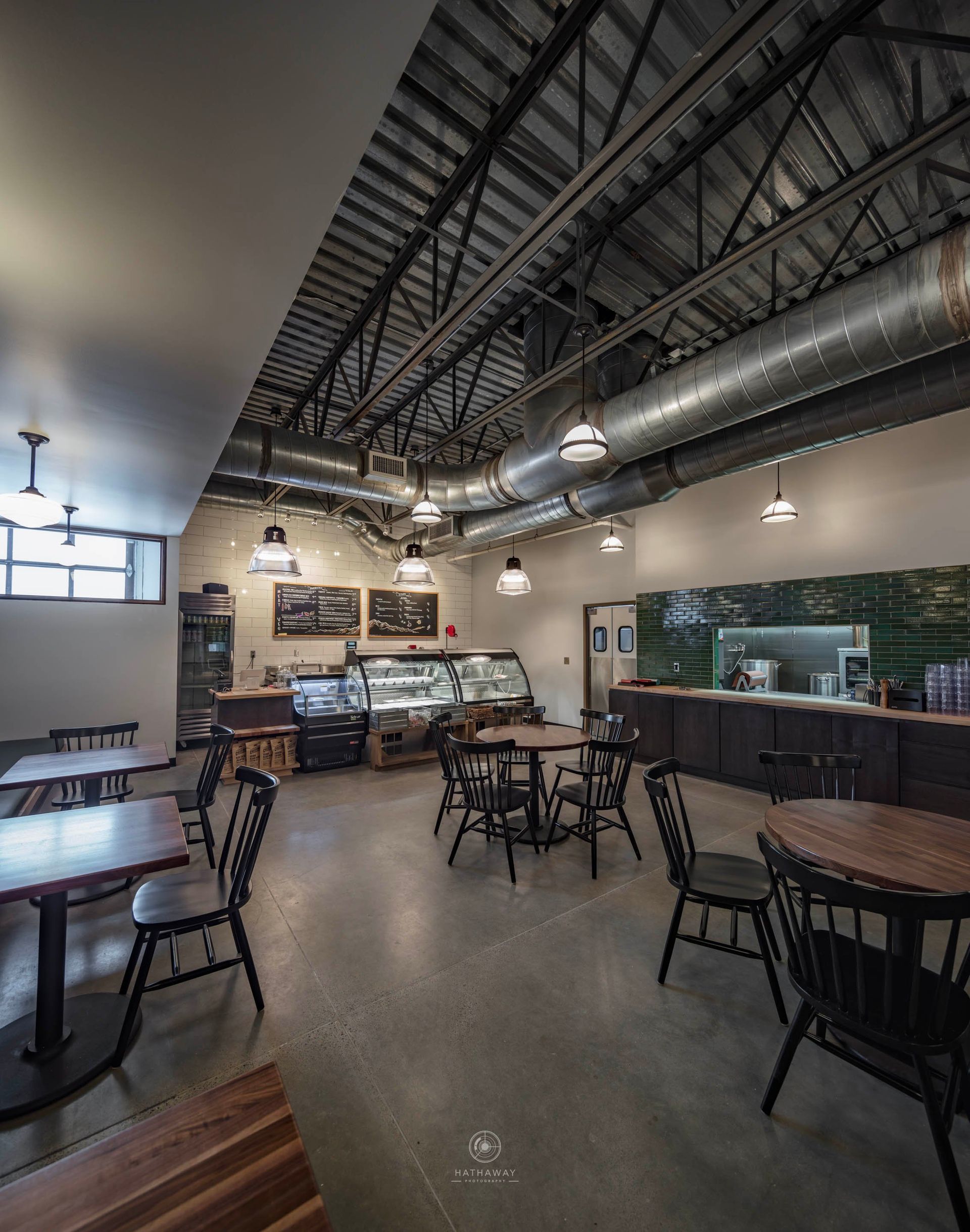 A modern cafe with an open industrial ceiling, concrete floors, wooden tables, and black chairs.