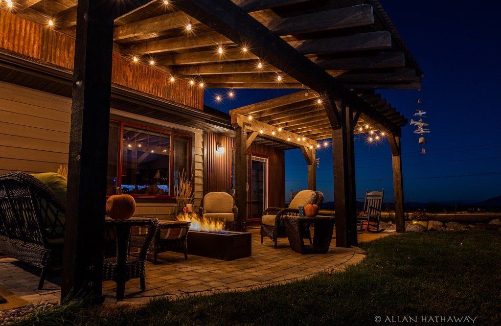 A cozy outdoor patio at twilight with string lights, a fire pit, and comfortable seating under a wooden pergola.