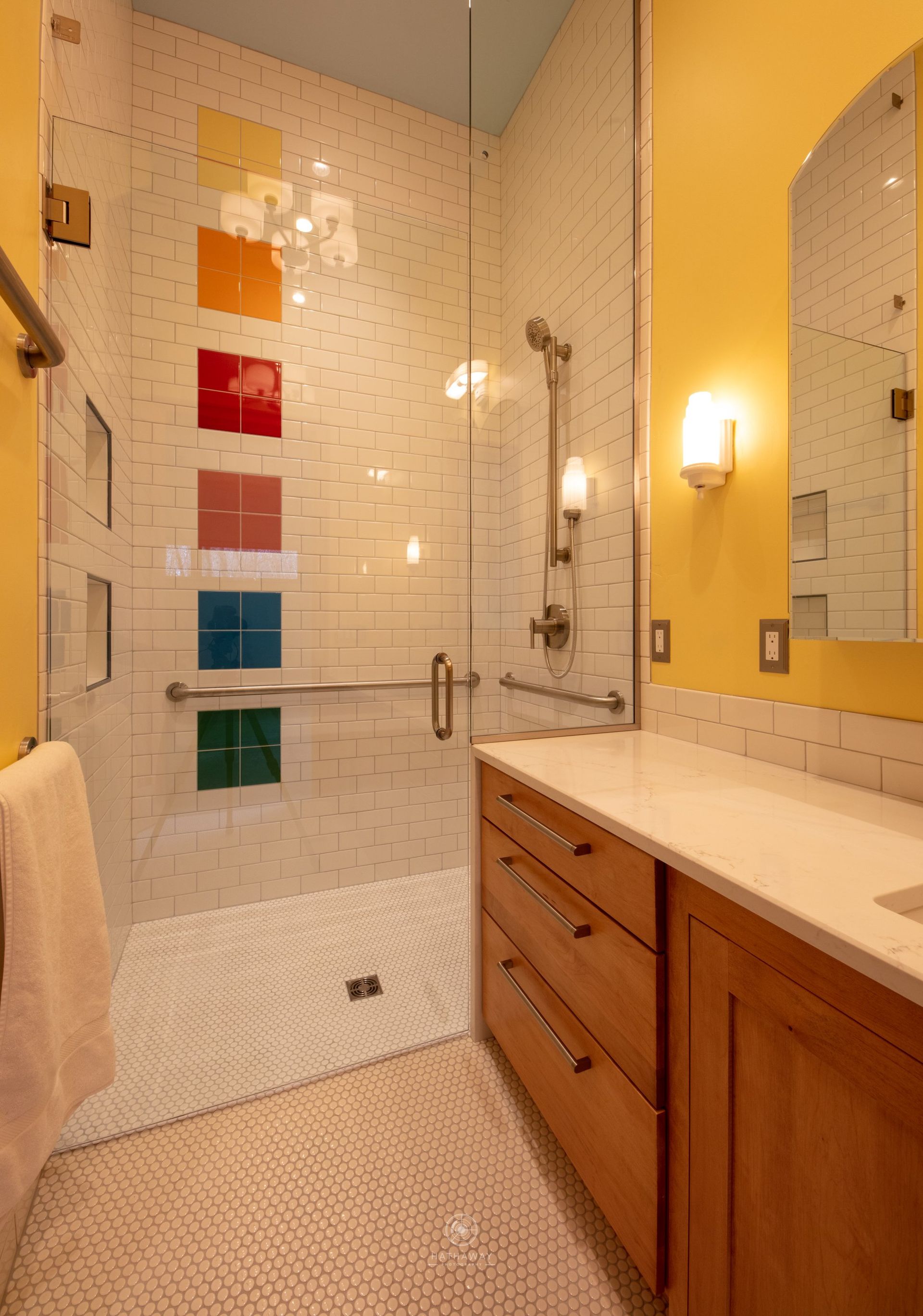 Modern bathroom with a shower featuring a vertical column of colorful tiles, a vanity, and a yellow accent wall.