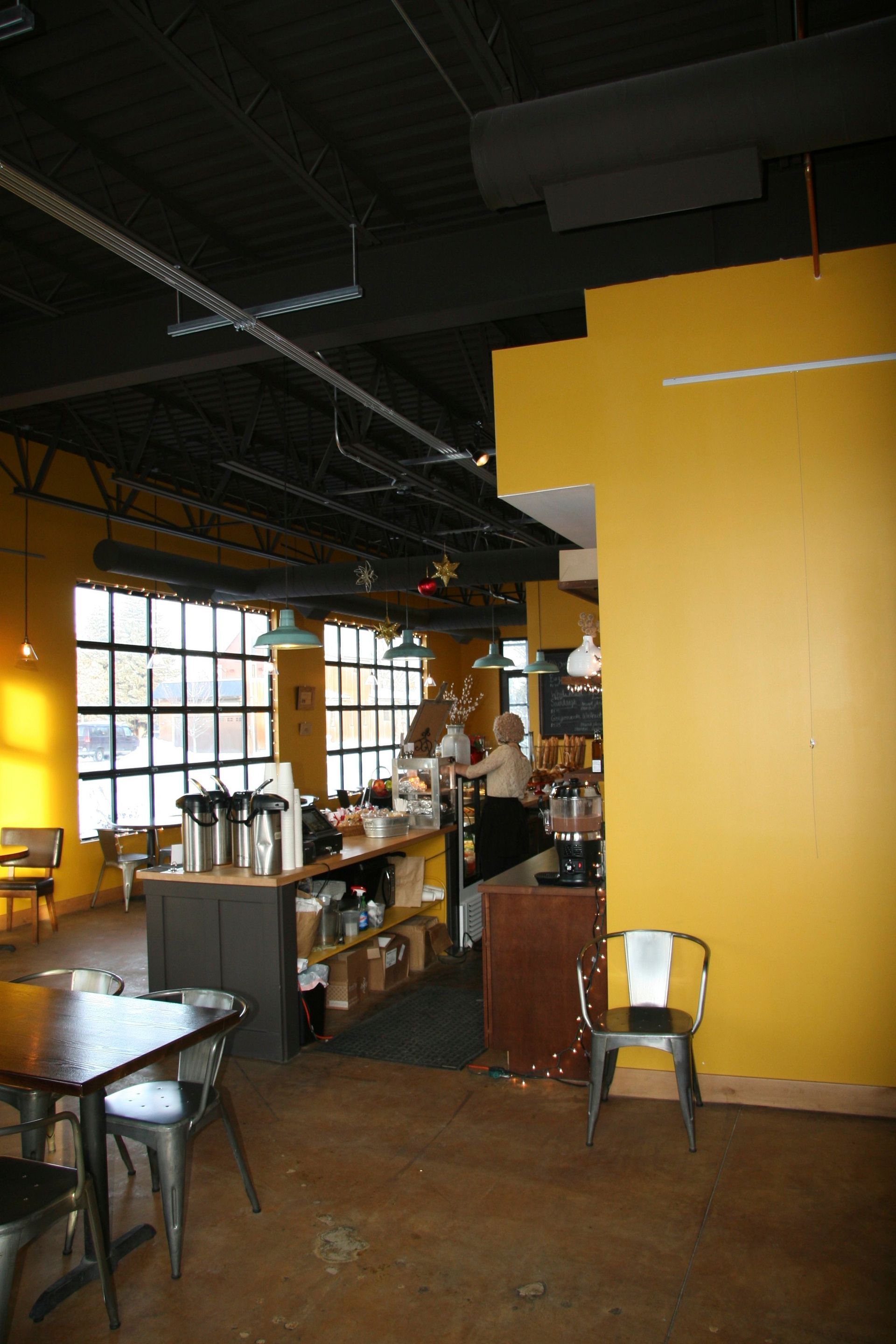 Interior of a coffee shop with dark ceilings, yellow walls, metal chairs, and a service counter facing large windows.