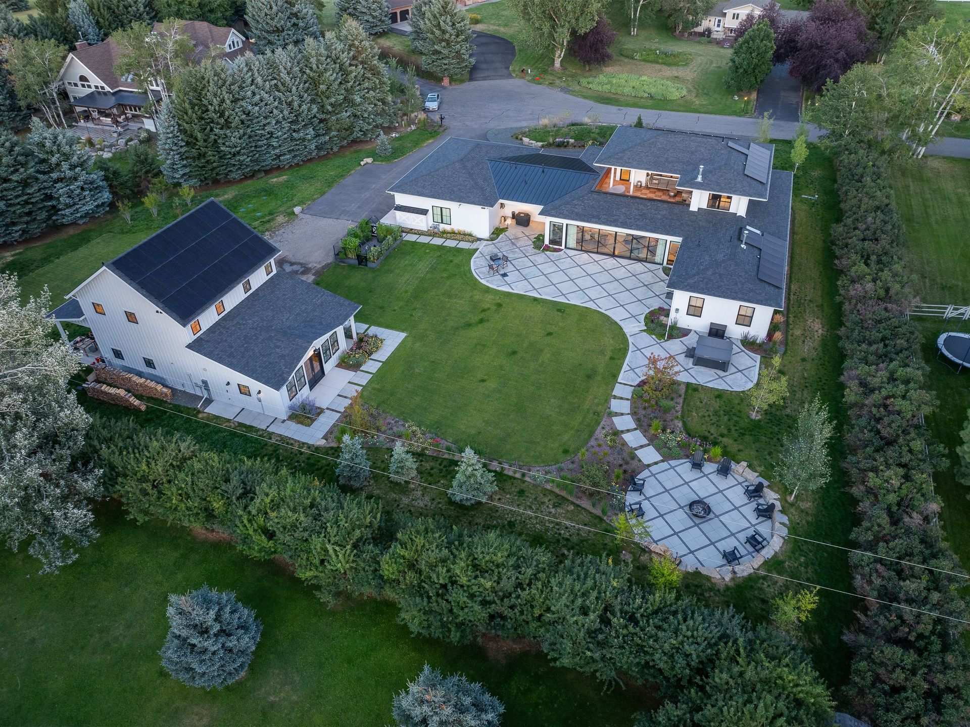 An aerial view of a white home with a dark roof, a detached garage, a large green lawn, and a stone patio with a fire pit.
