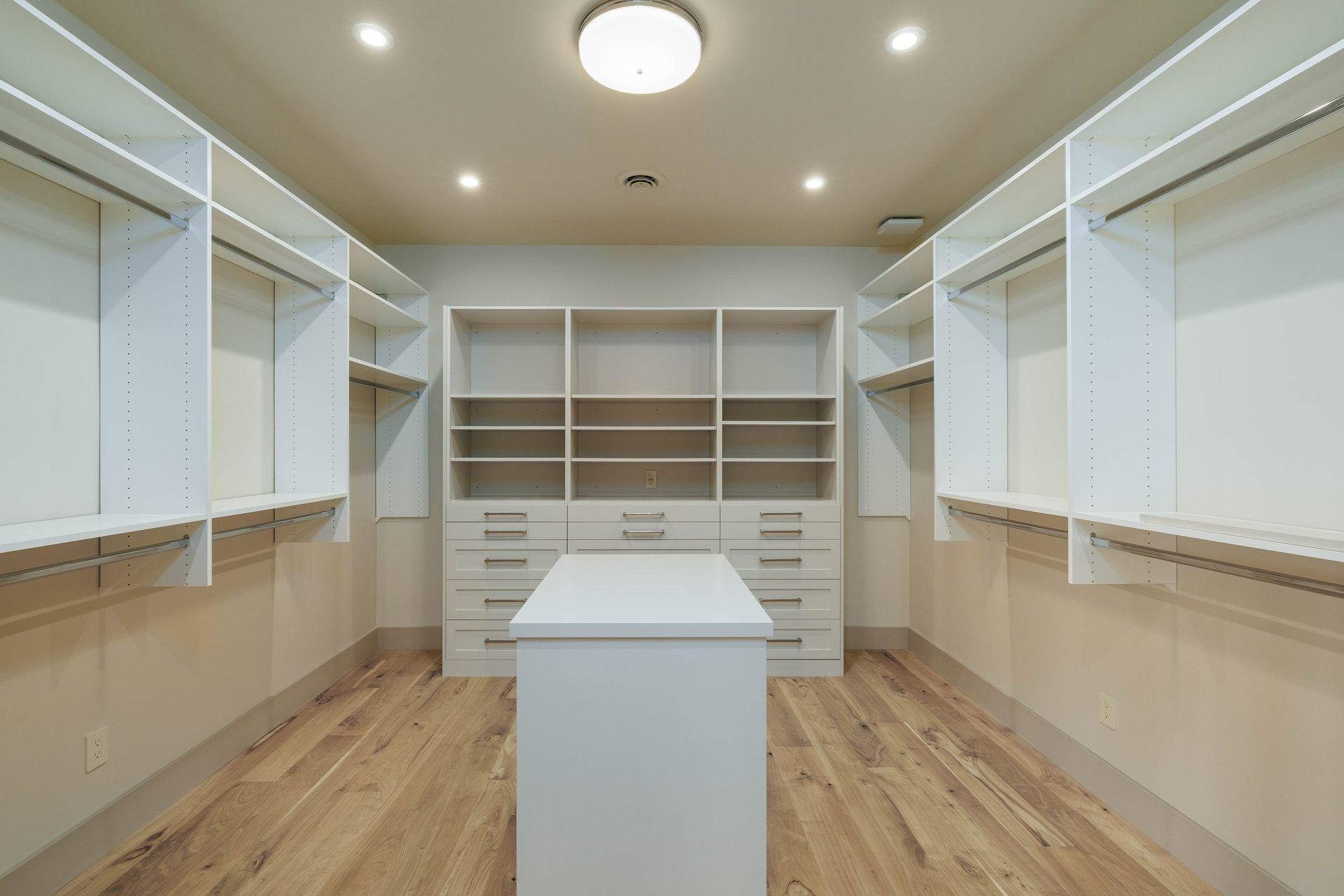 A walk-in closet featuring white shelving, a center island with drawers, and light wood flooring under recessed lighting.