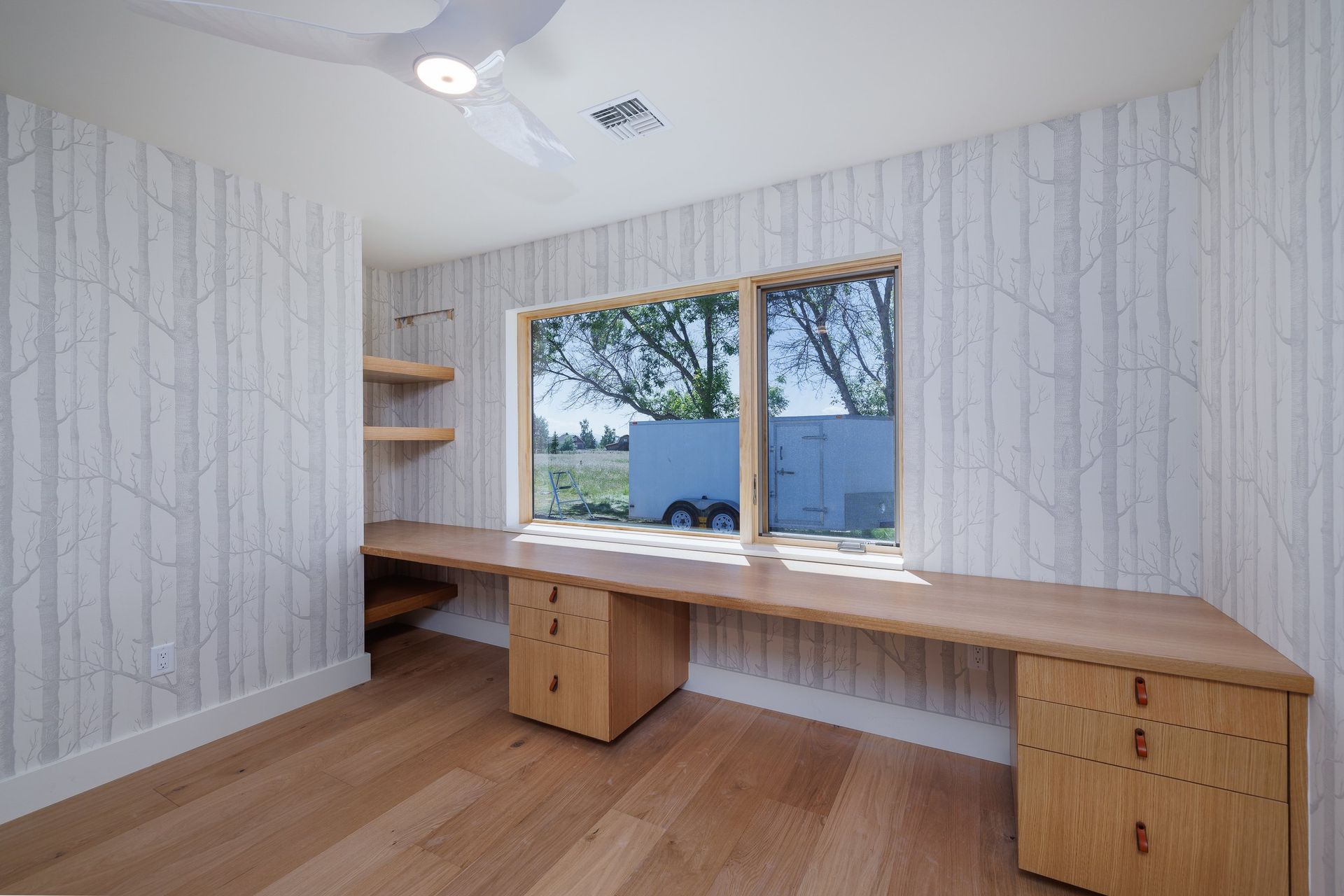 A home office features birch-patterned wallpaper, a long wooden desk with two drawer units, and a large window.