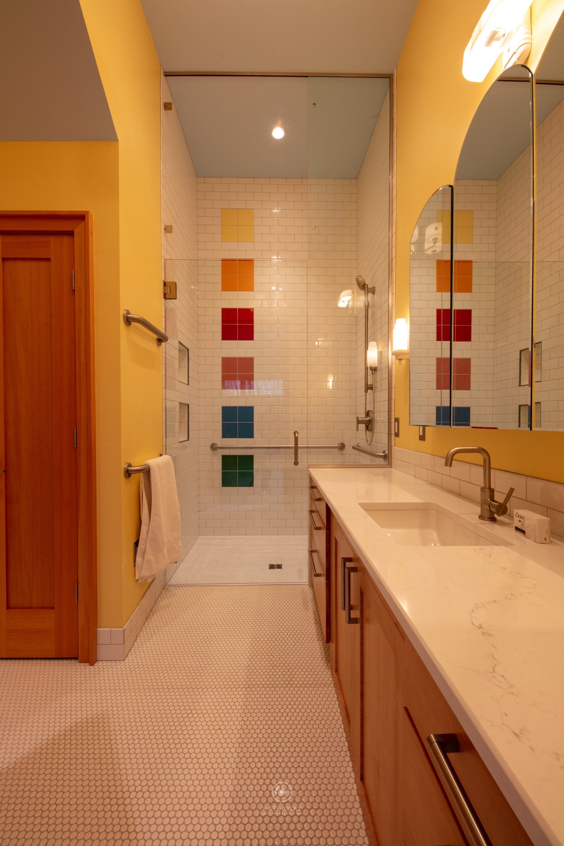 A bathroom with yellow walls, a wooden vanity, and a glass-enclosed shower featuring a column of colorful square tiles.