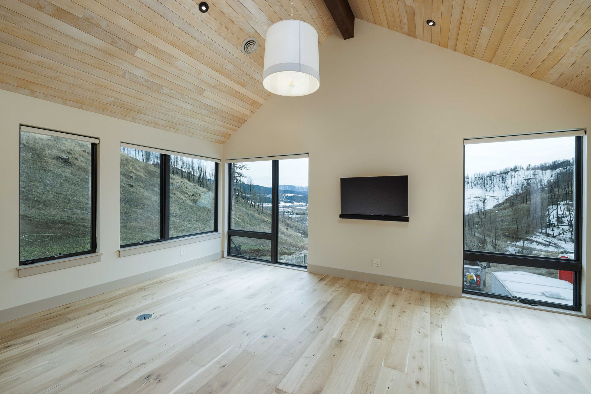 A spacious room with light hardwood floors, vaulted wood ceiling, a hanging pendant light, and windows overlooking hills.
