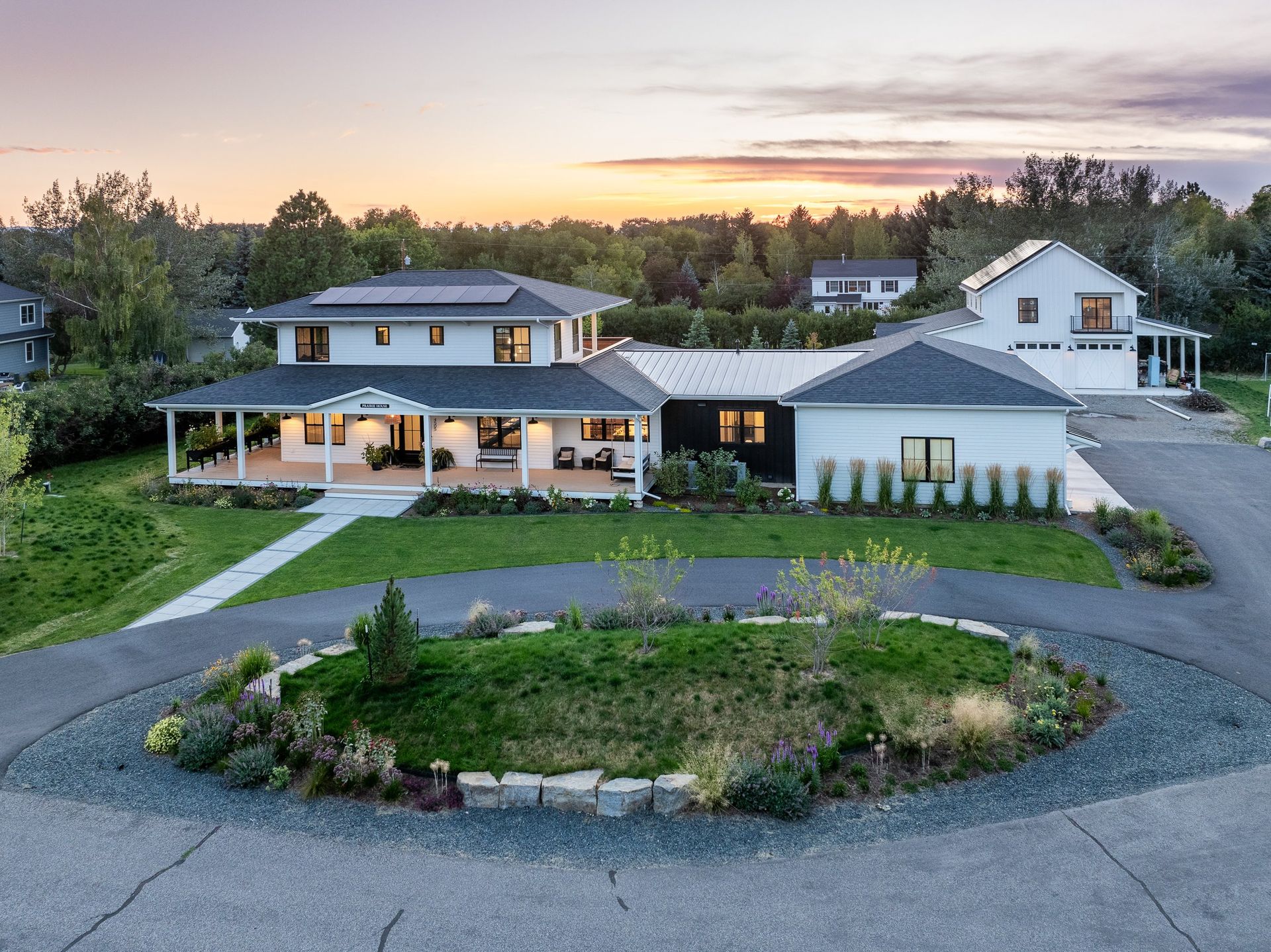 A white farmhouse-style home at sunset with a wrap-around porch, a detached garage, and a landscaped circular driveway.
