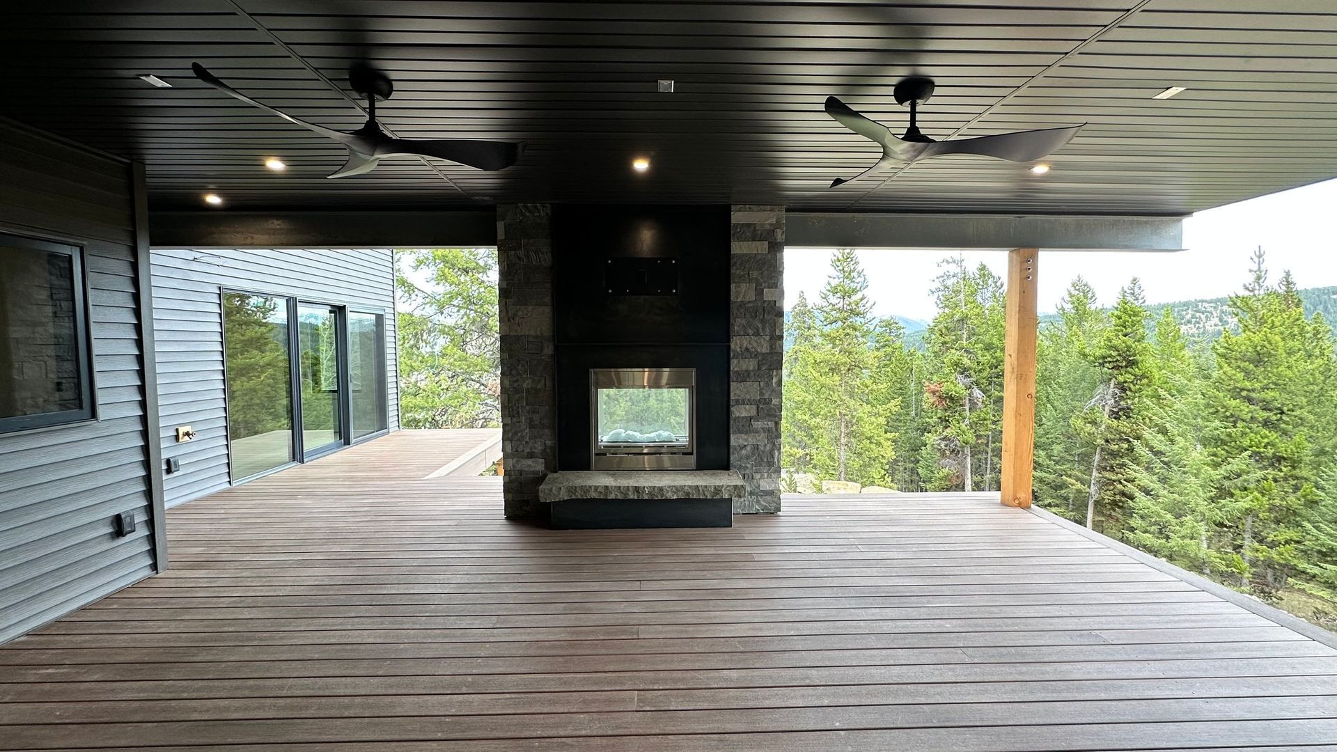 A modern covered outdoor deck with a central stone fireplace, wooden flooring, two ceiling fans, and a forest view.