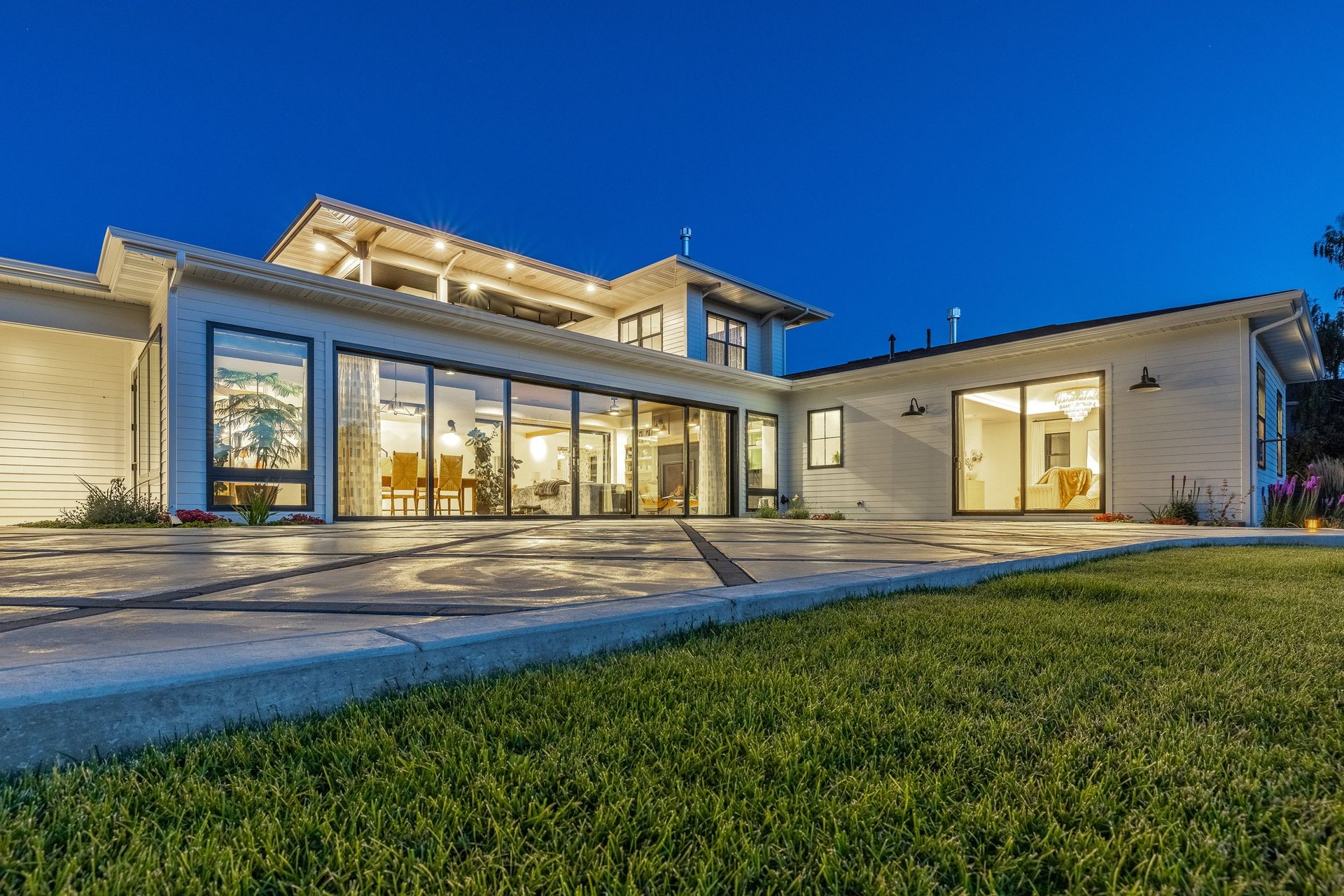 A modern white house with large glass sliding doors and exterior lighting at dusk, set behind a paved patio and lawn.