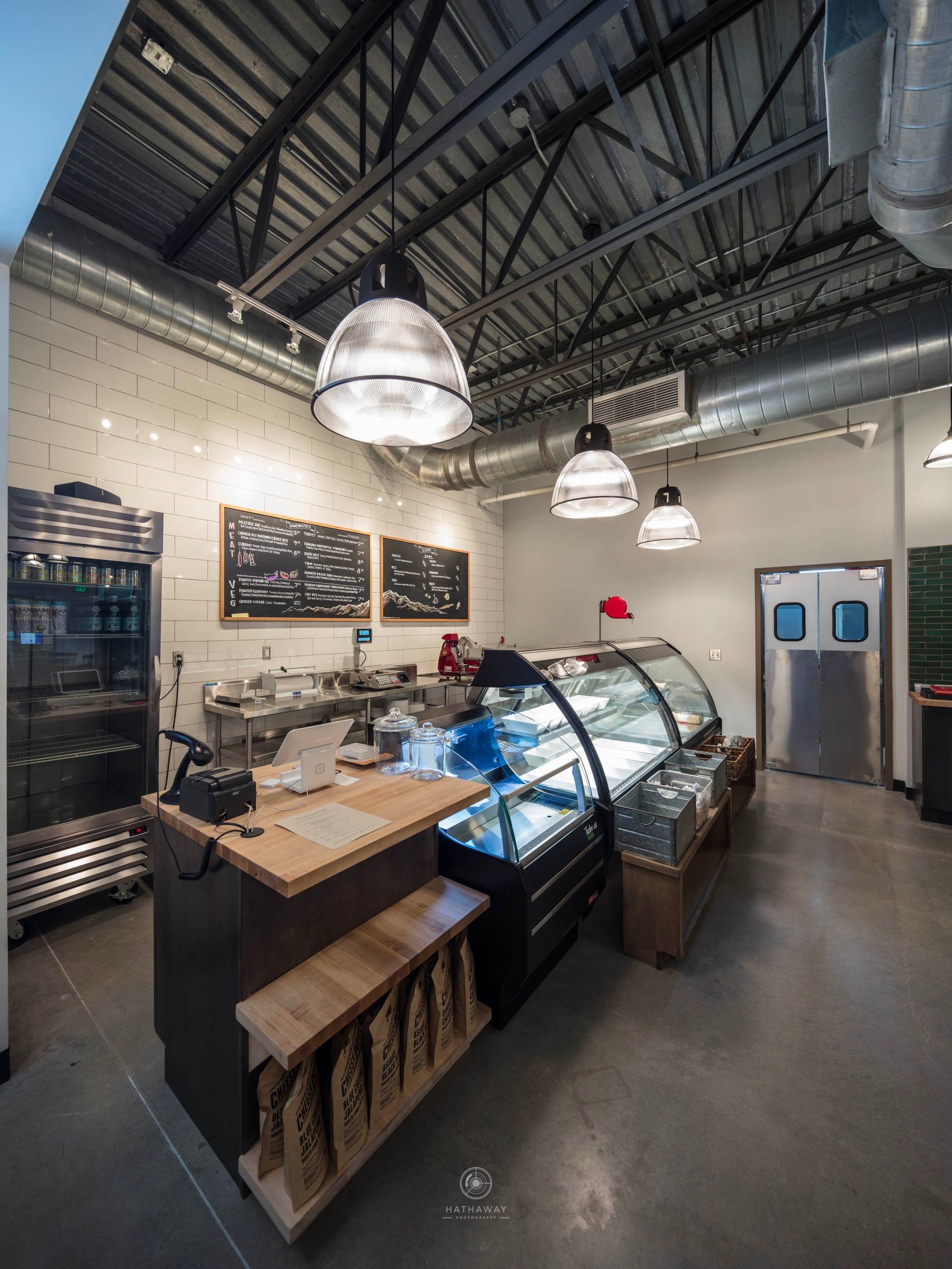 Interior view of a modern cafe counter with an industrial ceiling, display cases, menu boards, and a standing cooler.