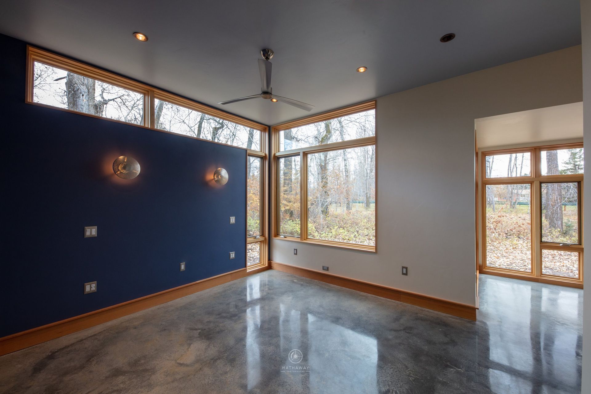 A room with polished concrete floors, a dark blue accent wall, a light gray wall, and large windows looking onto woods.