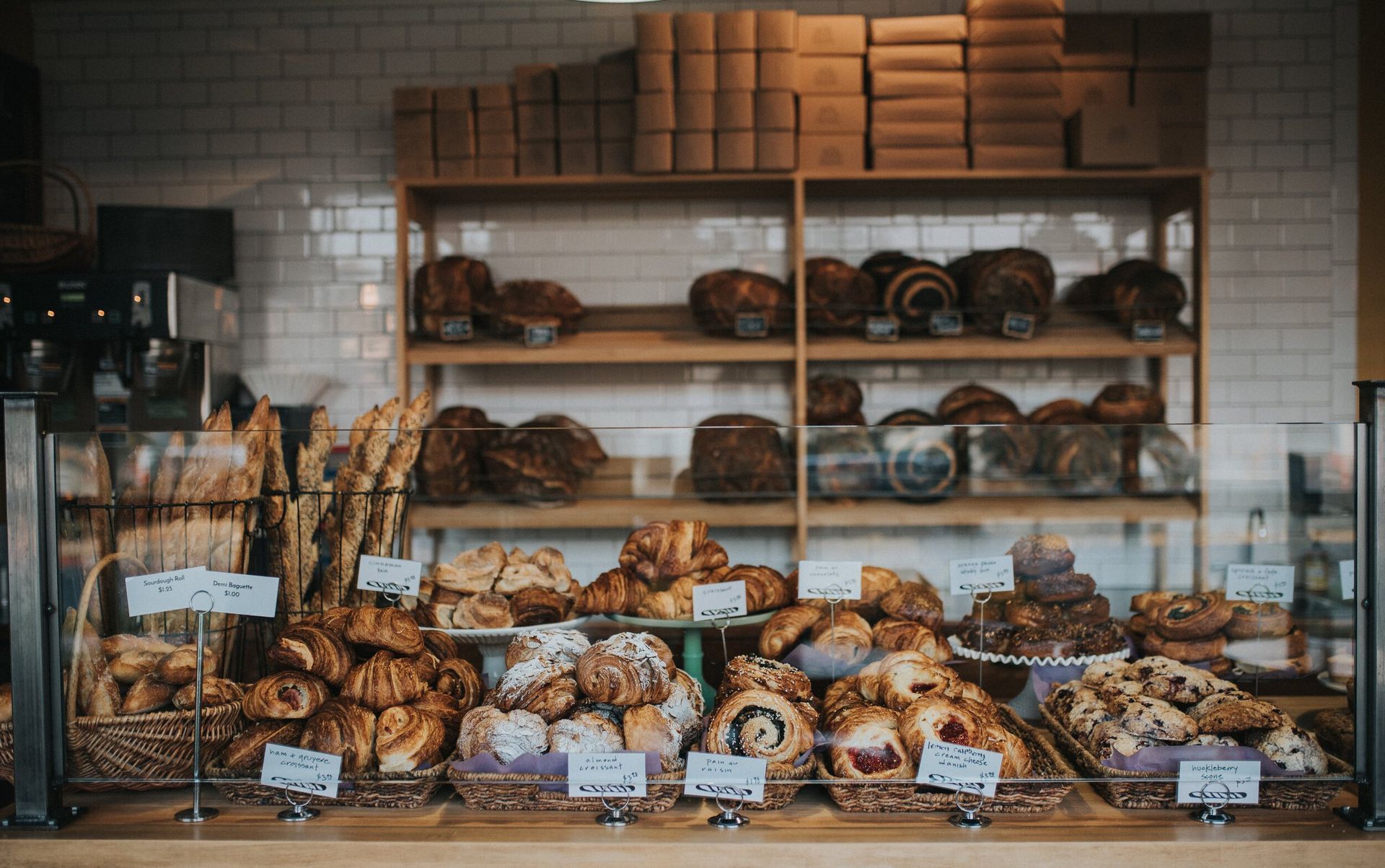 A display case filled with fresh croissants and pastries at a bakery, with loaves of bread on shelves in the background.
