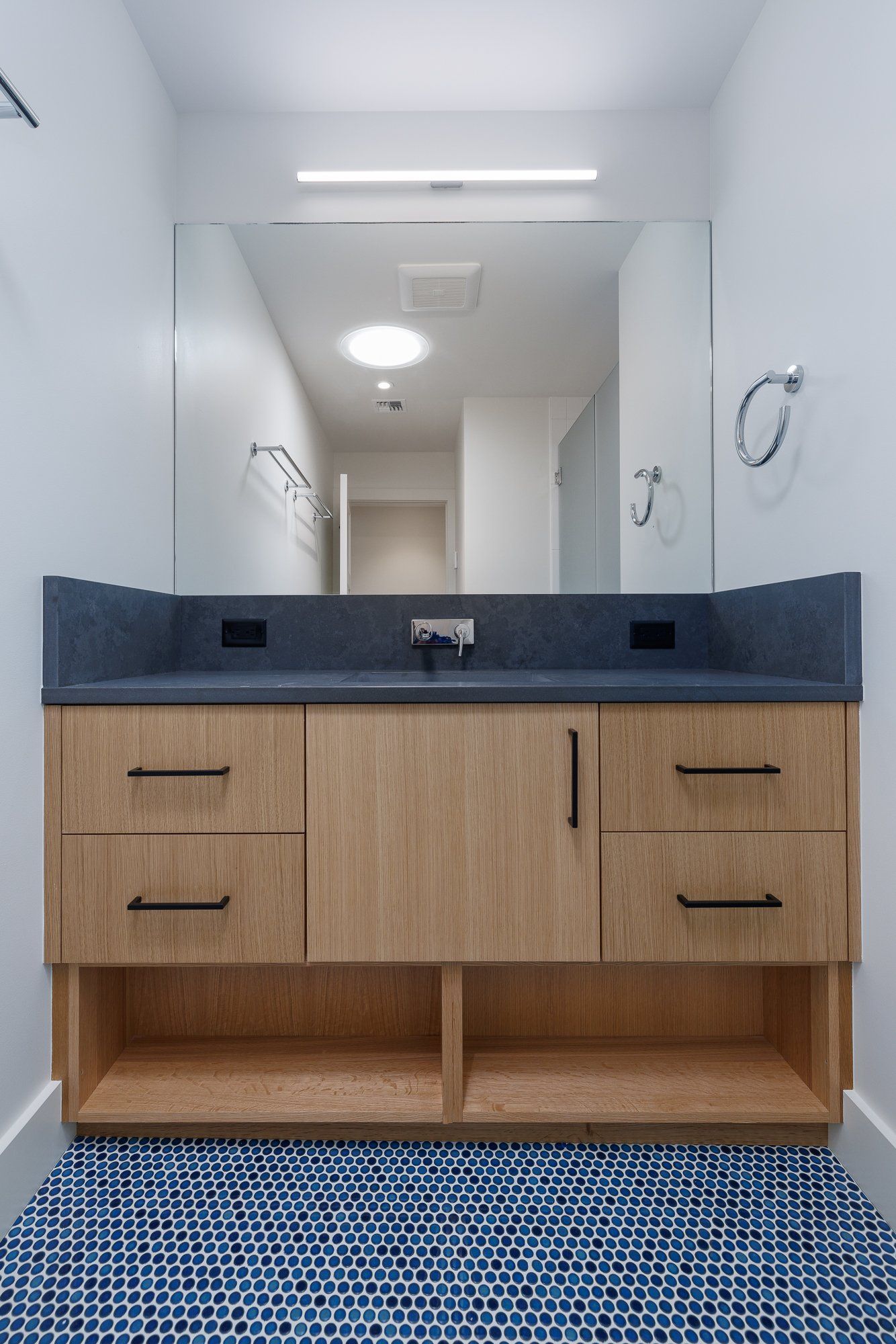 A modern bathroom vanity with wood-grain cabinets, a gray countertop, a large mirror, and blue patterned floor tiles.