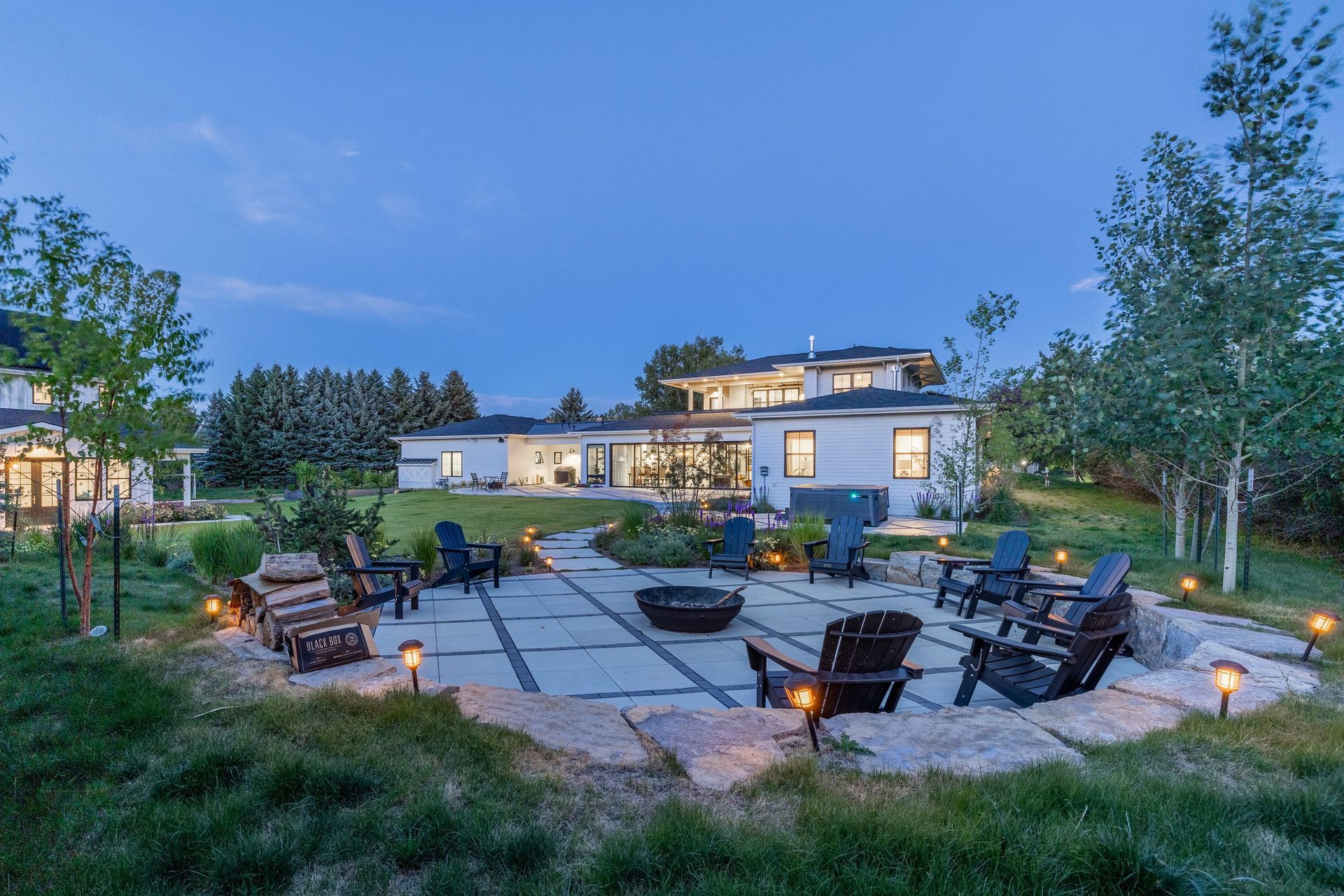 A stone patio with a fire pit and Adirondack chairs set in a grassy backyard at dusk, with a large modern house behind.