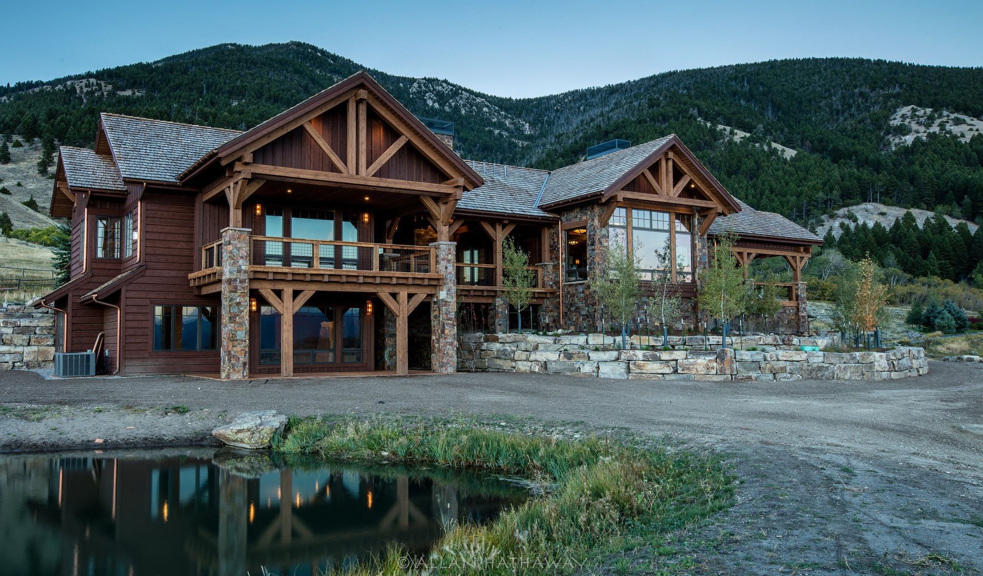 A rustic, multi-level wood and stone mountain home with a large deck sits beside a small pond under a clear sky.