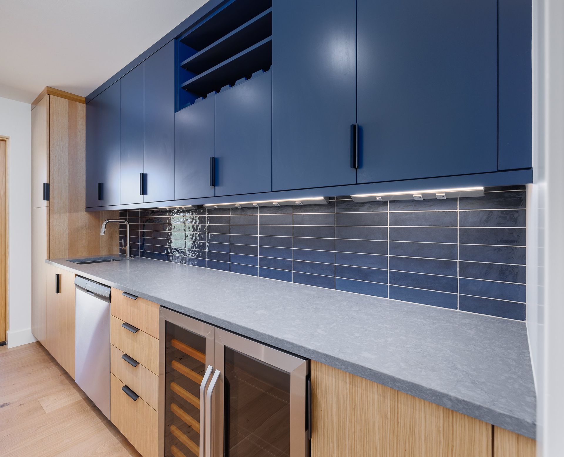 Modern kitchen featuring blue upper cabinets, light wood lower cabinets, gray stone countertops, and a striped tile backsplash.