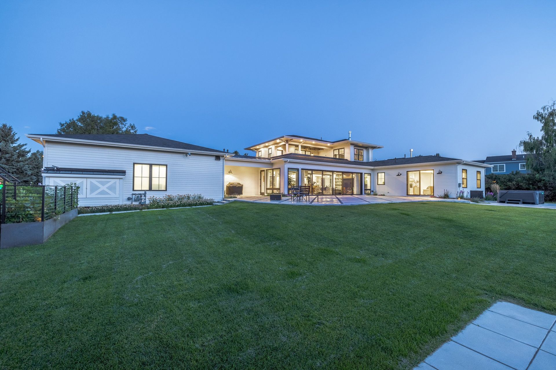 A multi-level contemporary home with warm interior lighting illuminates a backyard lawn at dusk under a clear blue sky.