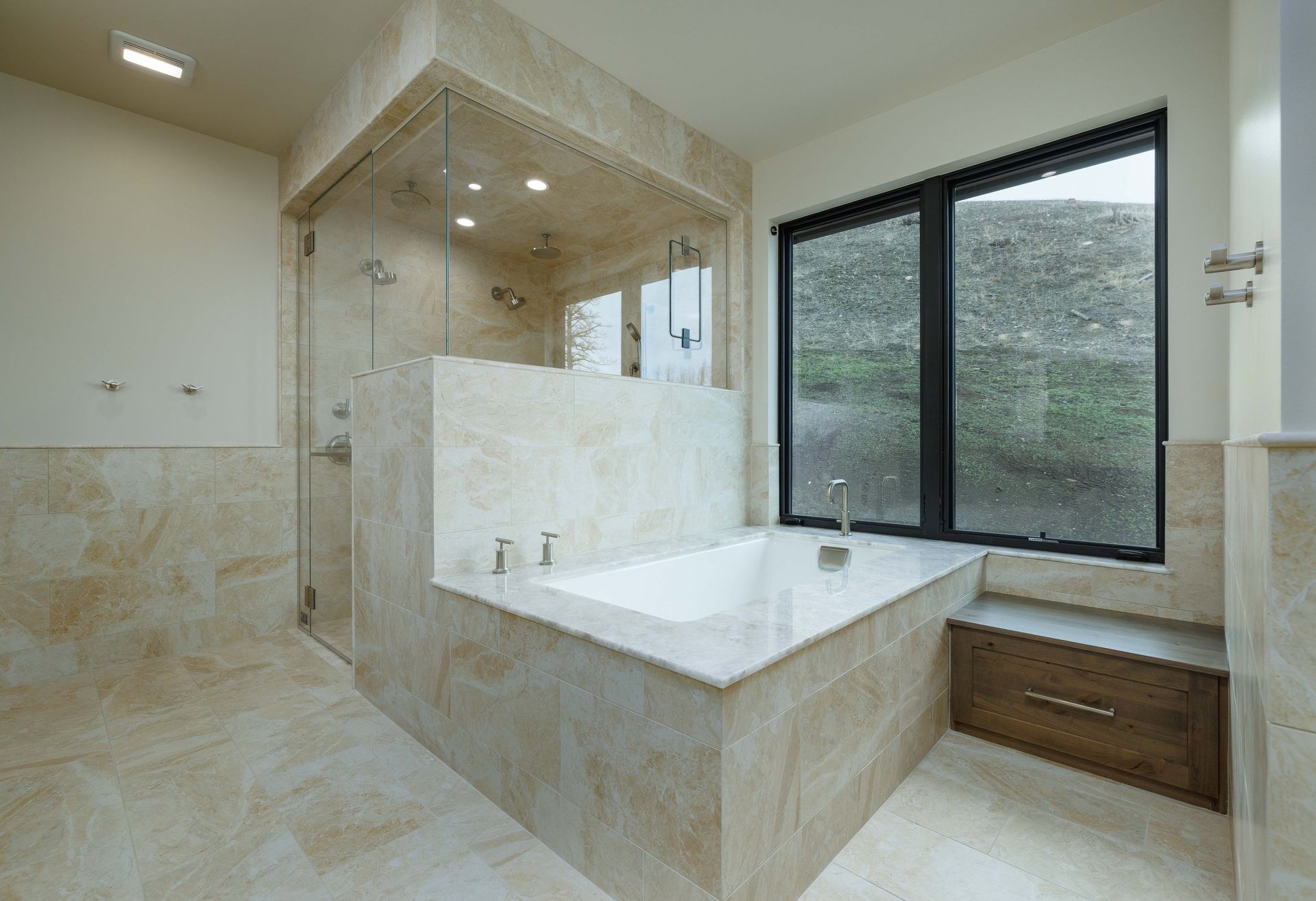 A luxurious bathroom with a glass-enclosed shower, stone-tiled bathtub, large window, and wooden storage drawer.