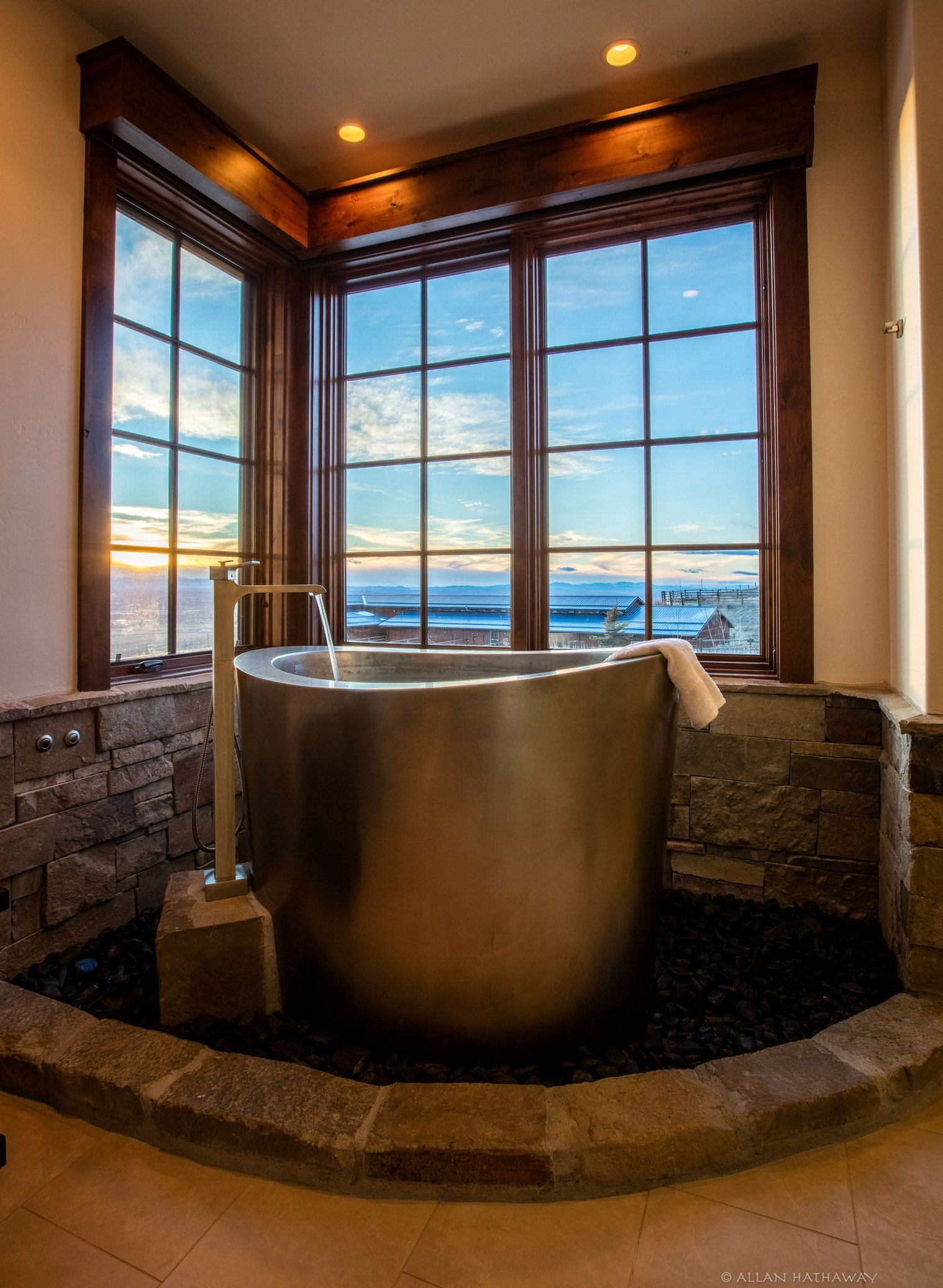 A circular metal bathtub sits in a stone-floored alcove featuring large windows with a view of a body of water at sunset.