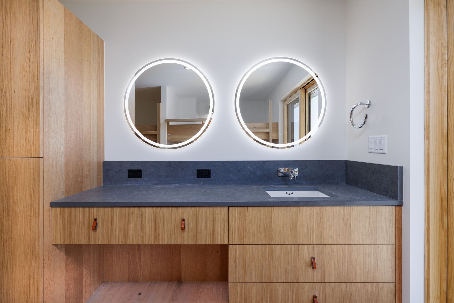 A modern bathroom vanity with wood cabinets, a dark gray stone countertop, and two illuminated circular mirrors.