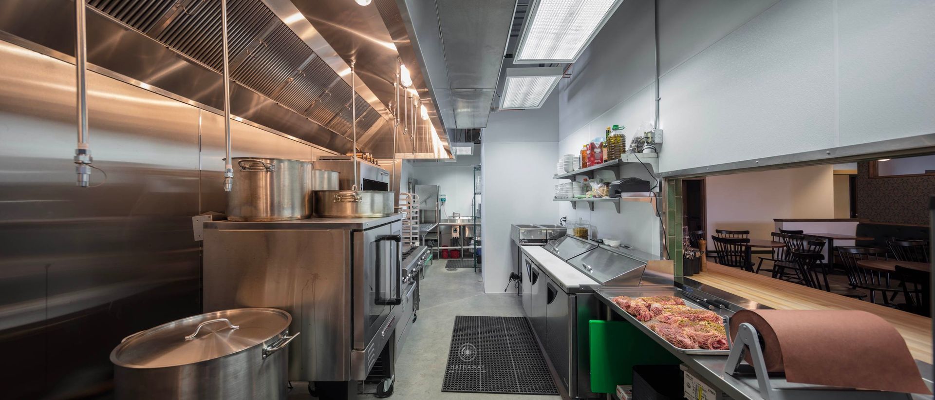 A commercial kitchen featuring stainless steel appliances, a large exhaust hood, prep tables, and a pass-through window.