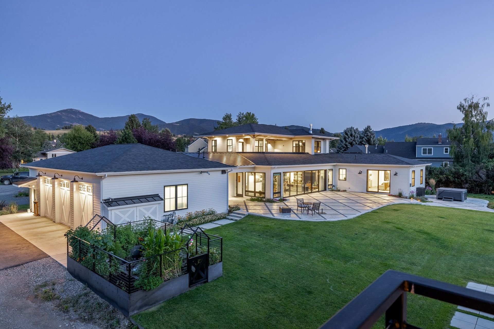 A modern two-story home at dusk with a three-car garage, a vegetable garden, and a patio overlooking a backyard lawn.