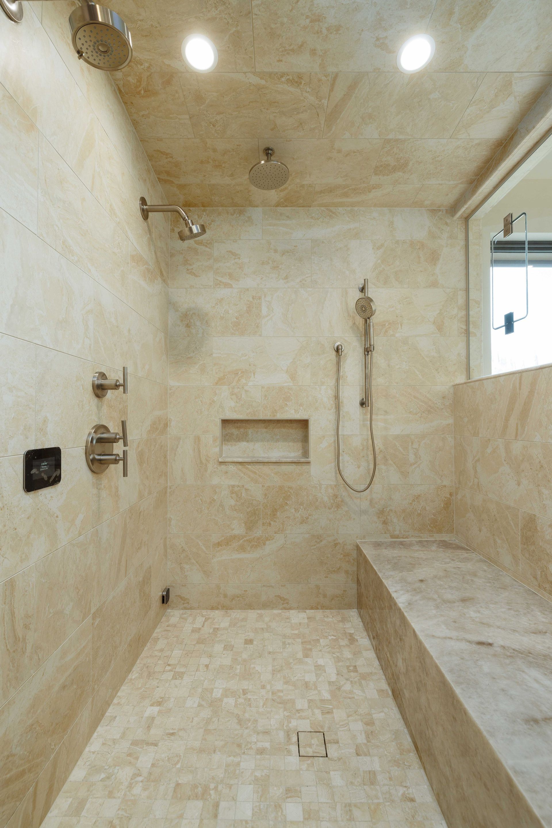 A walk-in shower with beige stone walls, a bench, a handheld showerhead, multiple overhead fixtures, and a recessed niche.