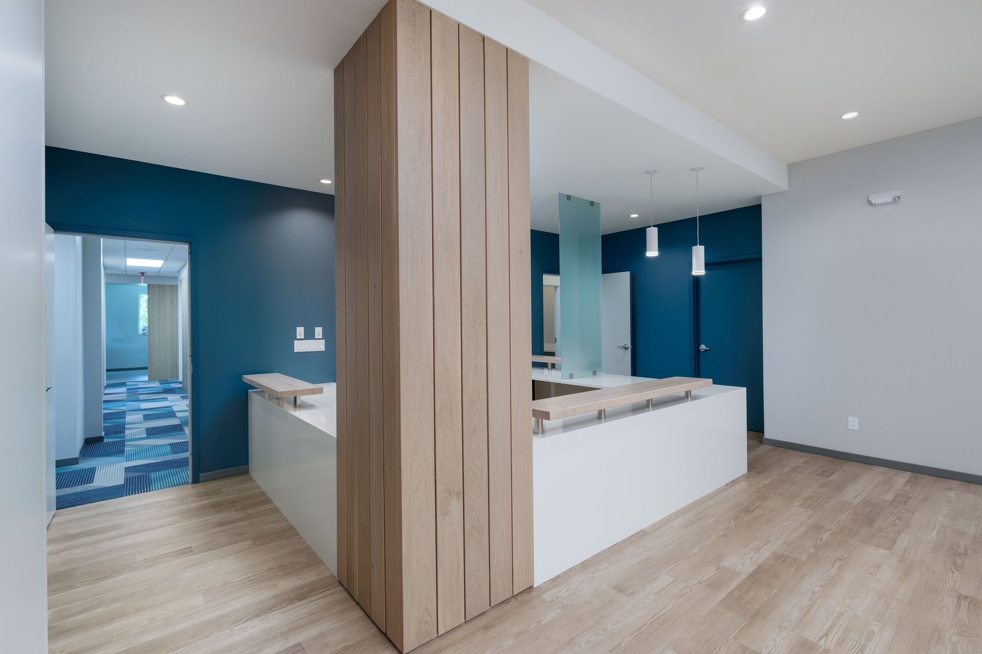 Modern office lobby with a central wood-slatted pillar, white reception desks, teal accent walls, and light wood flooring.