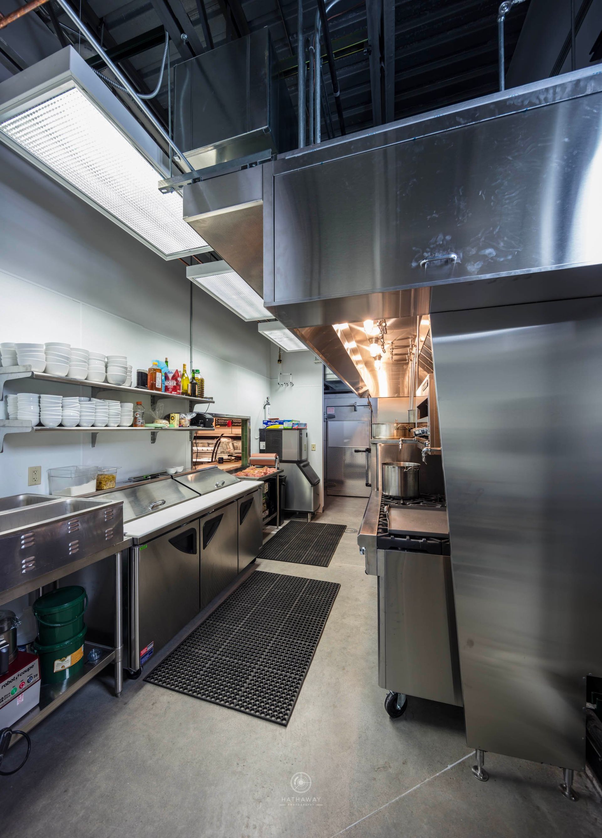 A commercial kitchen featuring stainless steel counters, a large ventilation hood, open shelving, and black floor mats.
