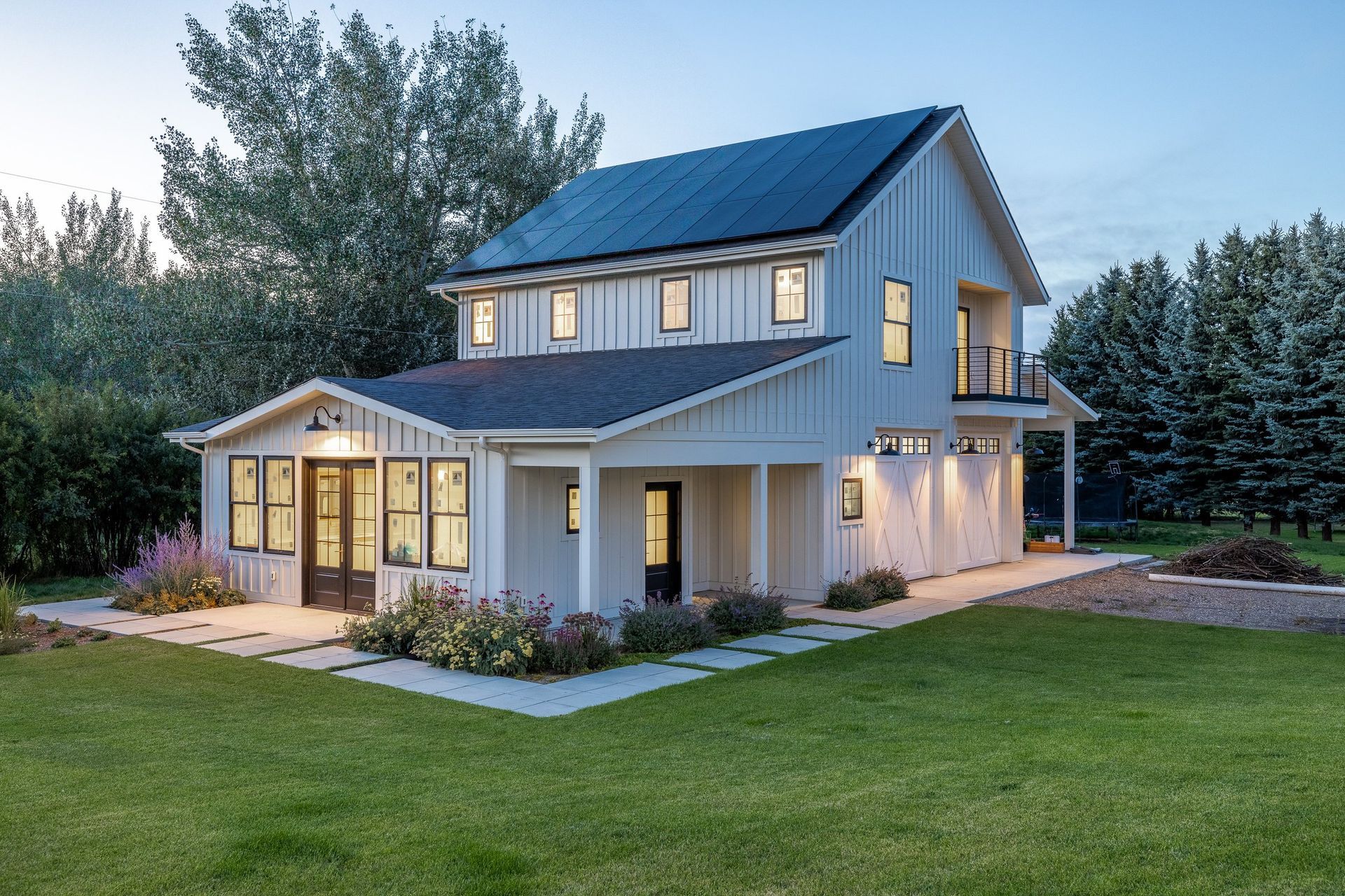 A white, modern two-story farmhouse with a dark roof and solar panels, surrounded by a green lawn and trees at dusk.