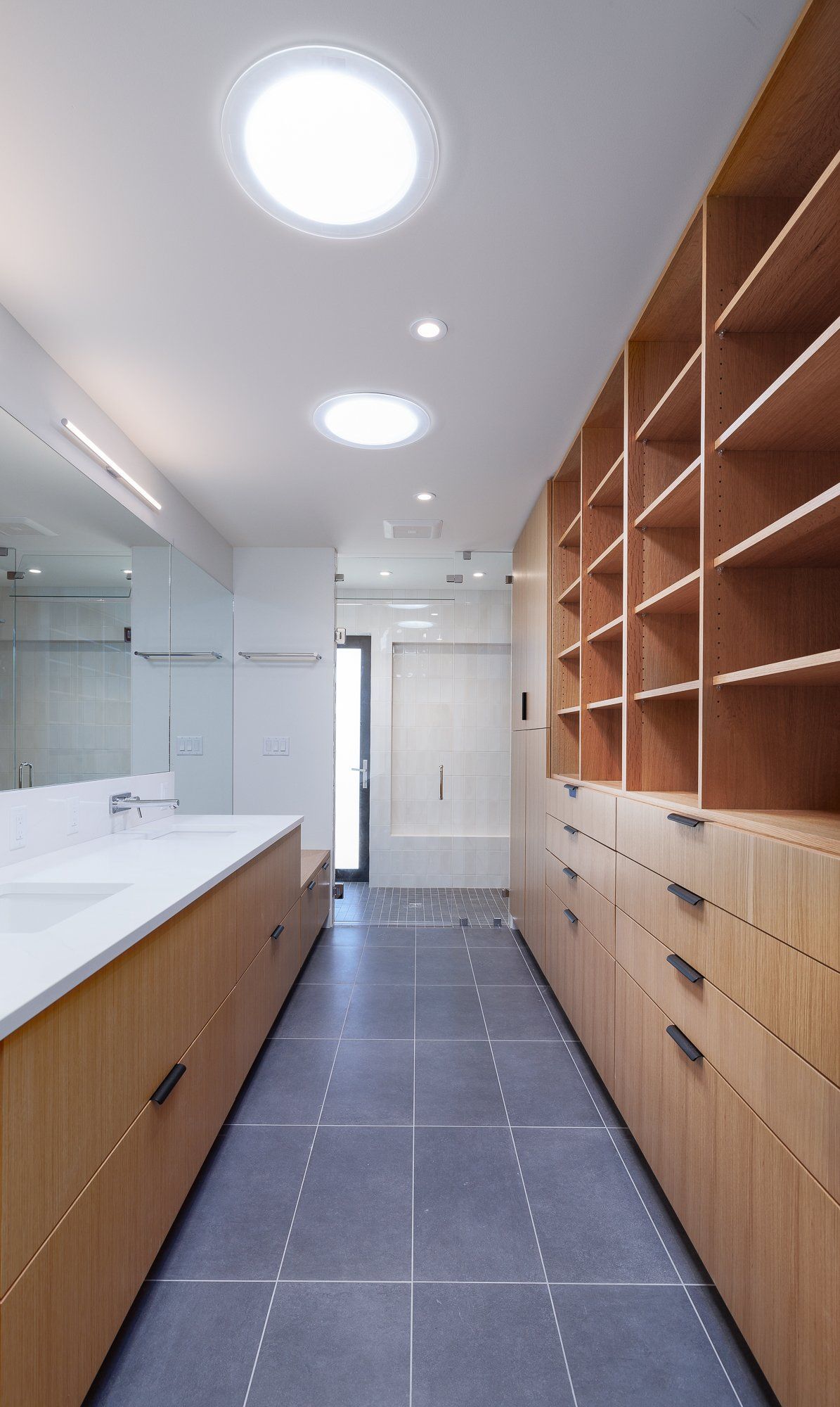 Modern bathroom with light wood cabinets, gray tile floors, white countertops, a large mirror, and two circular skylights.