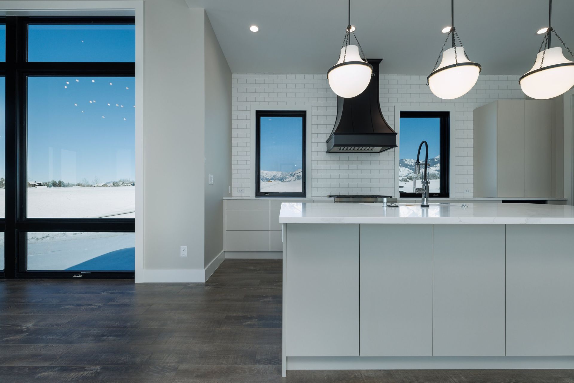 A modern, bright kitchen featuring a white island, light-colored cabinets, a black range hood, and large windows with snow.