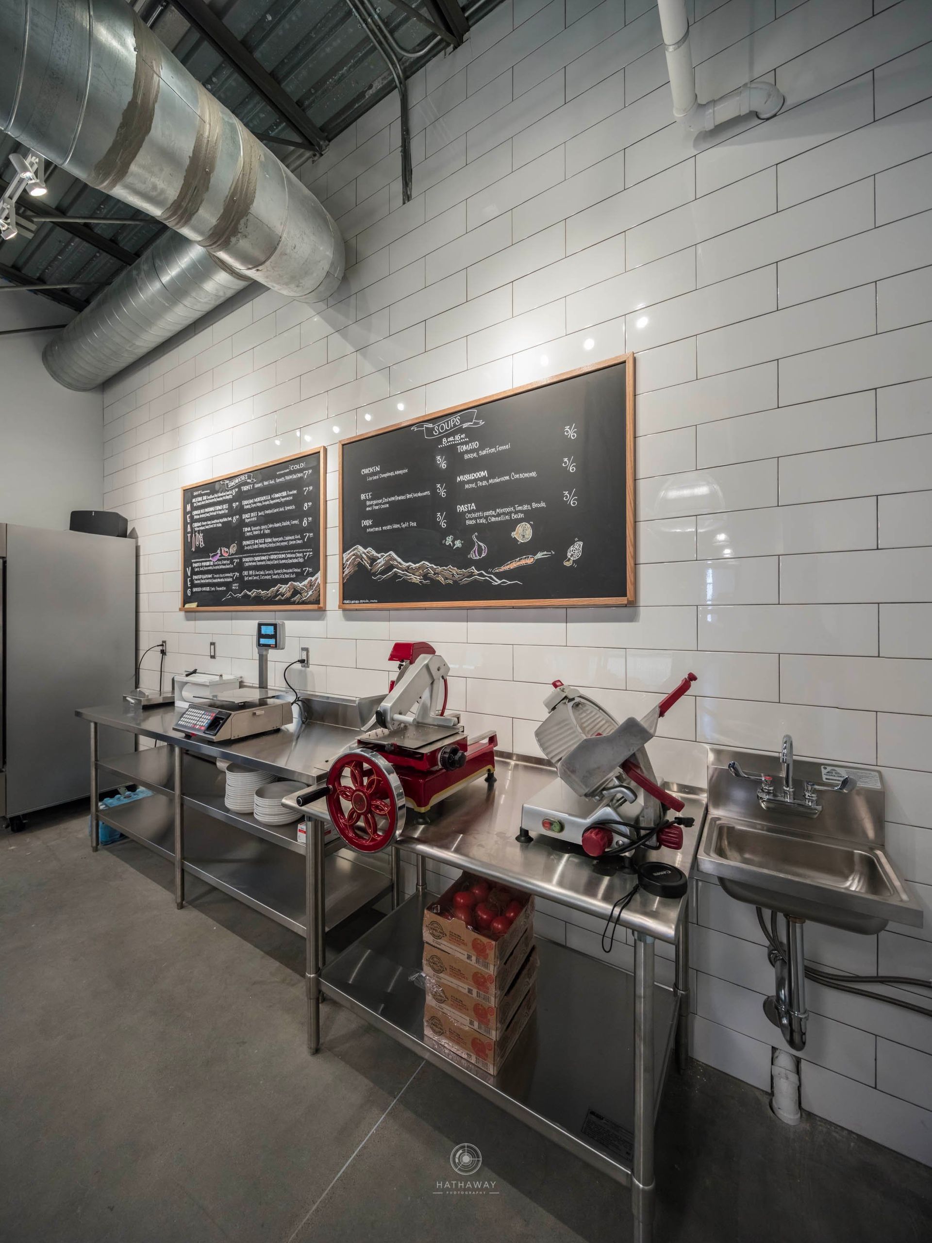 A commercial kitchen area with stainless steel prep tables, two meat slicers, a wall-mounted sink, and two menu chalkboards.