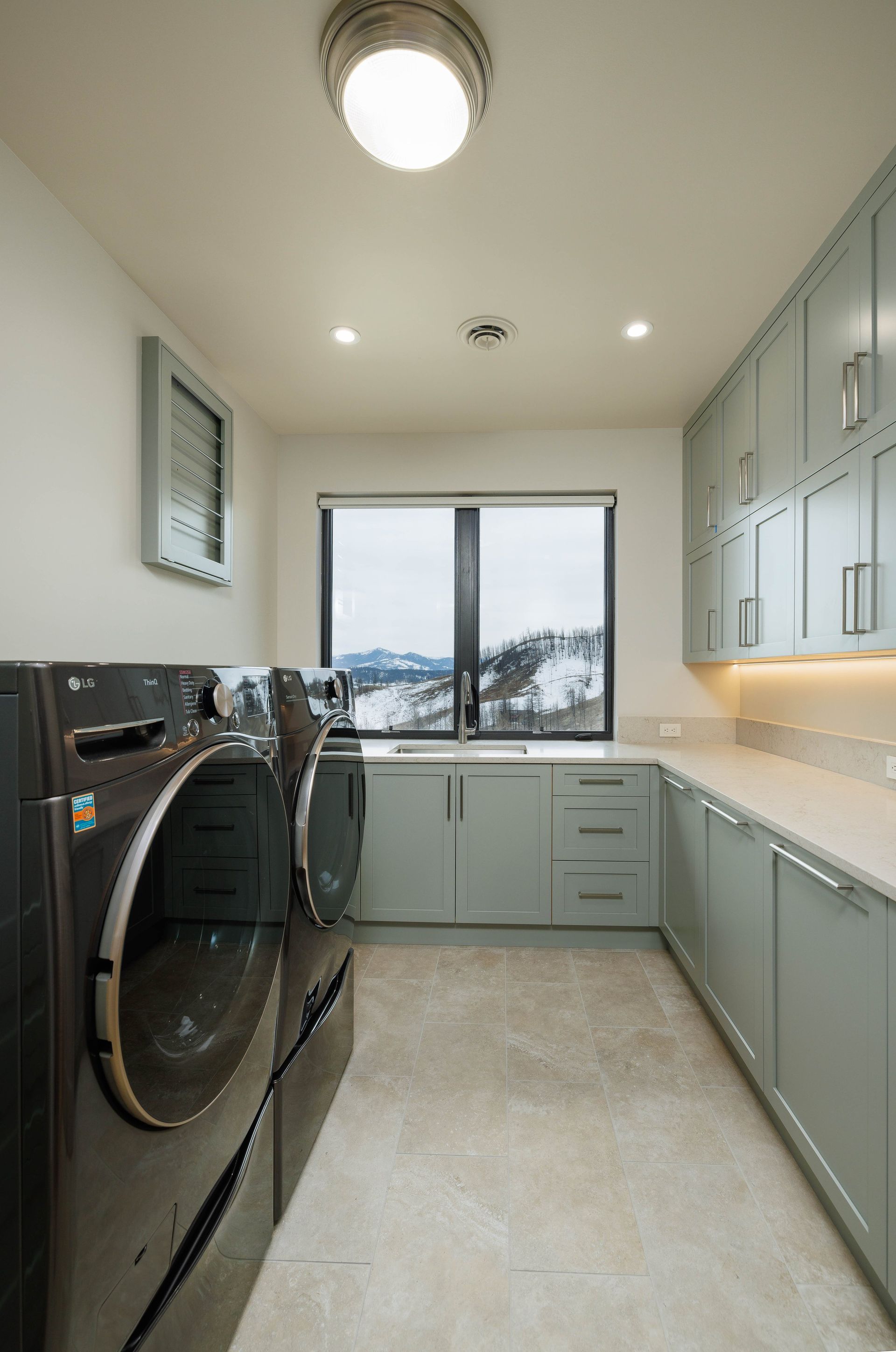 A laundry room with dark gray appliances, light green cabinetry, and a large window overlooking a snow-covered landscape.