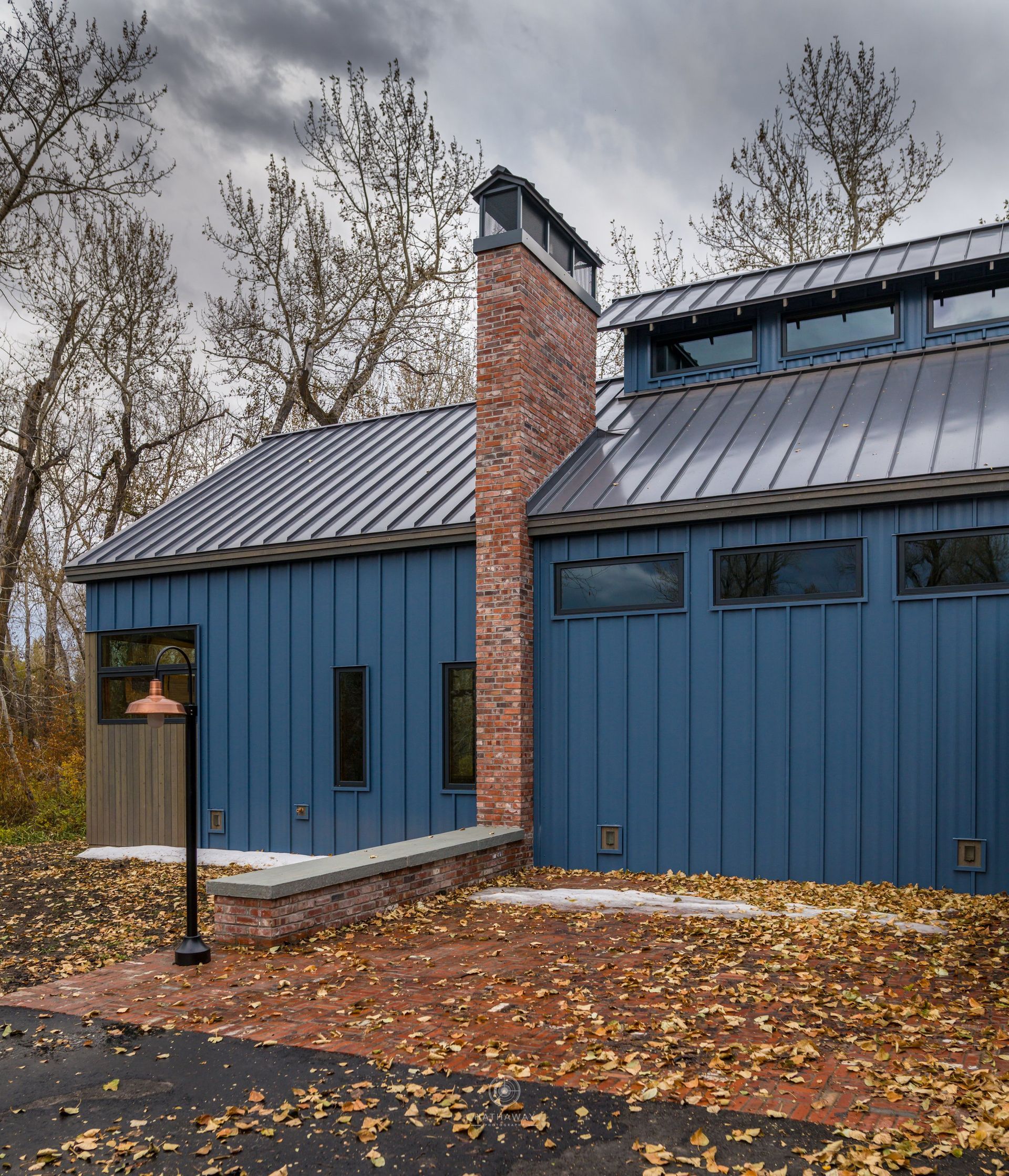 A blue vertical-siding house with a brick chimney and metal roof stands on a lot covered in autumn leaves.