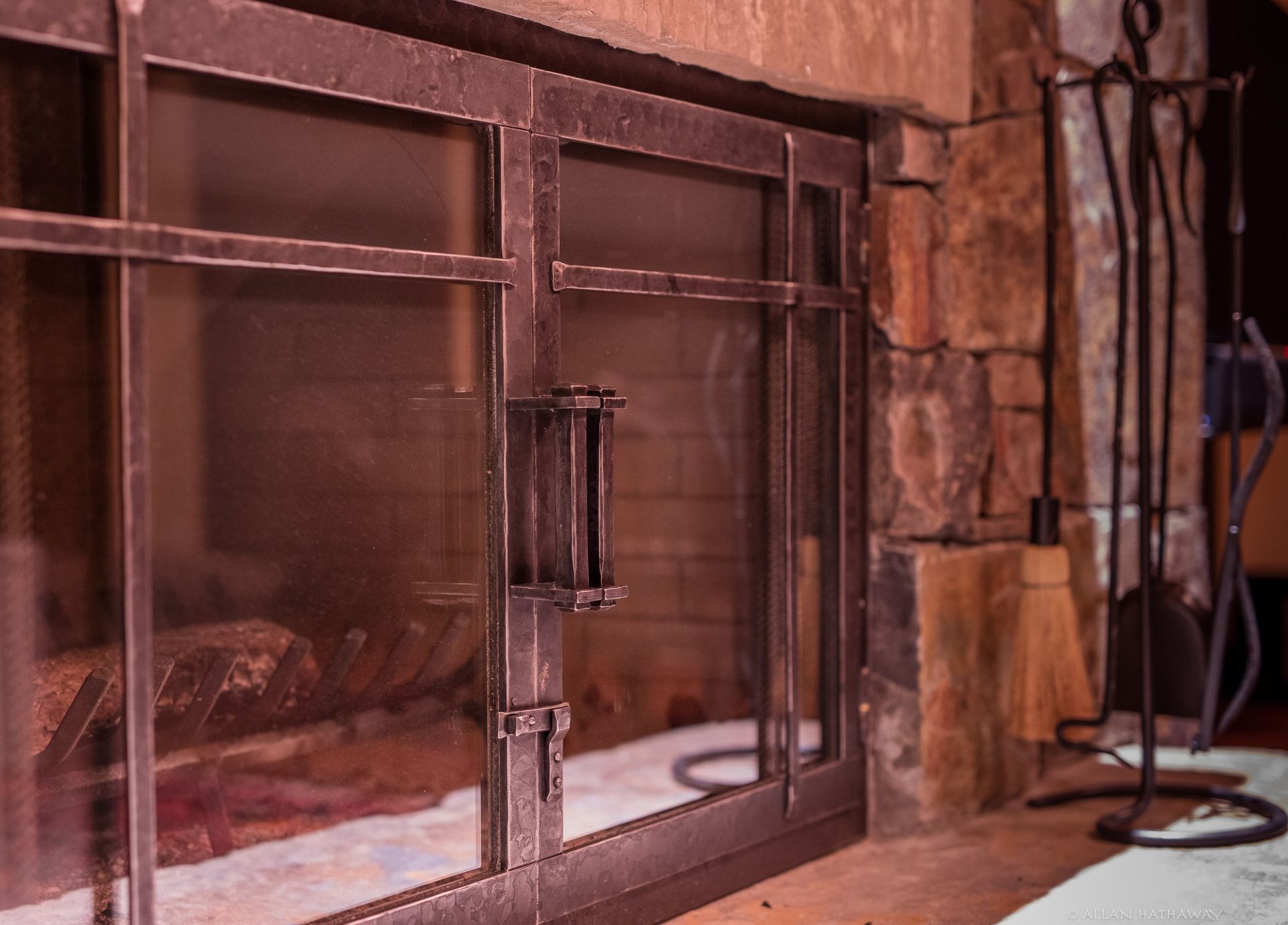 Close-up of a rustic metal fireplace door with glass panels, situated next to a set of fireplace tools on a stone hearth.