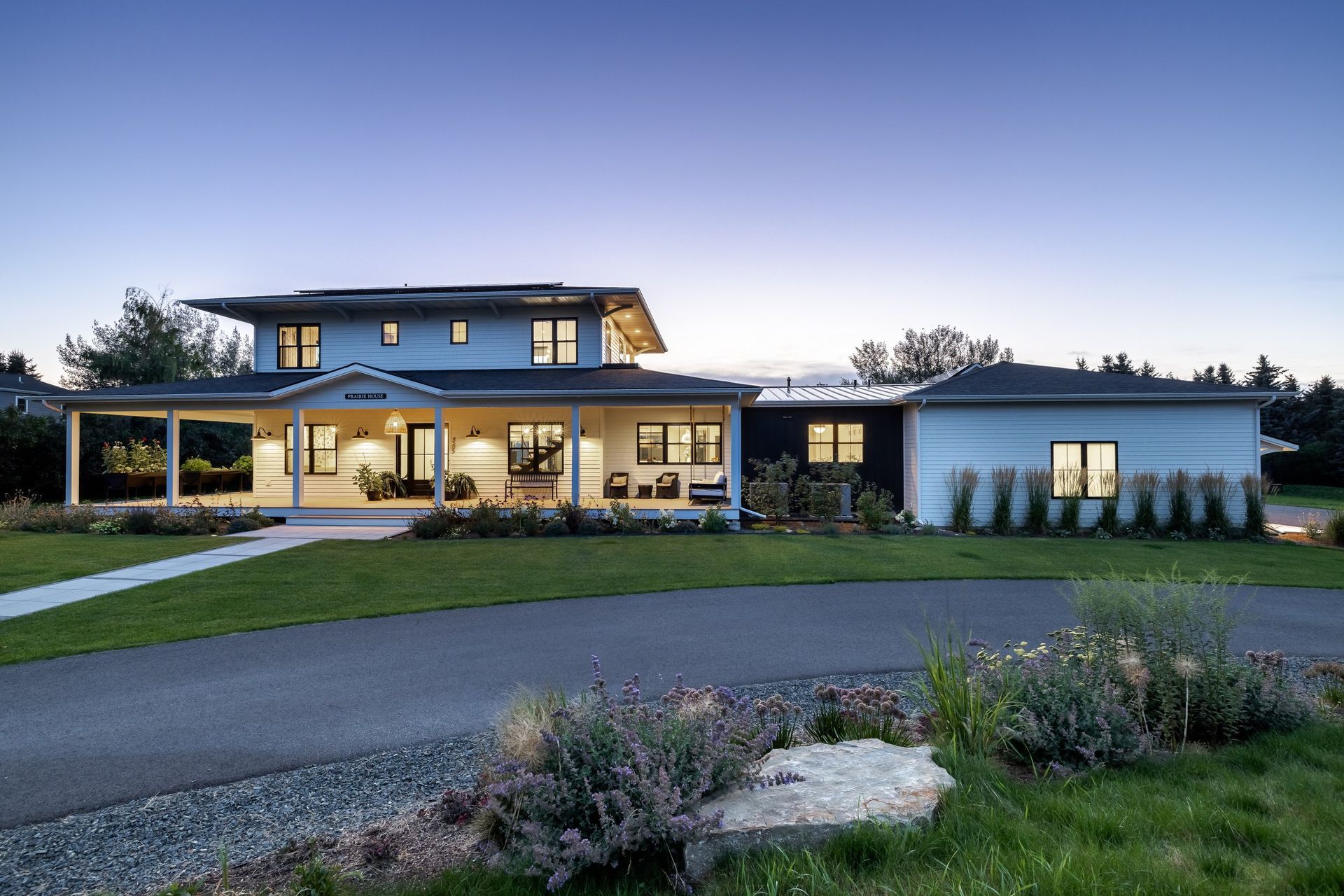 A two-story white modern farmhouse with a wide porch and attached garage, surrounded by a lawn at twilight.