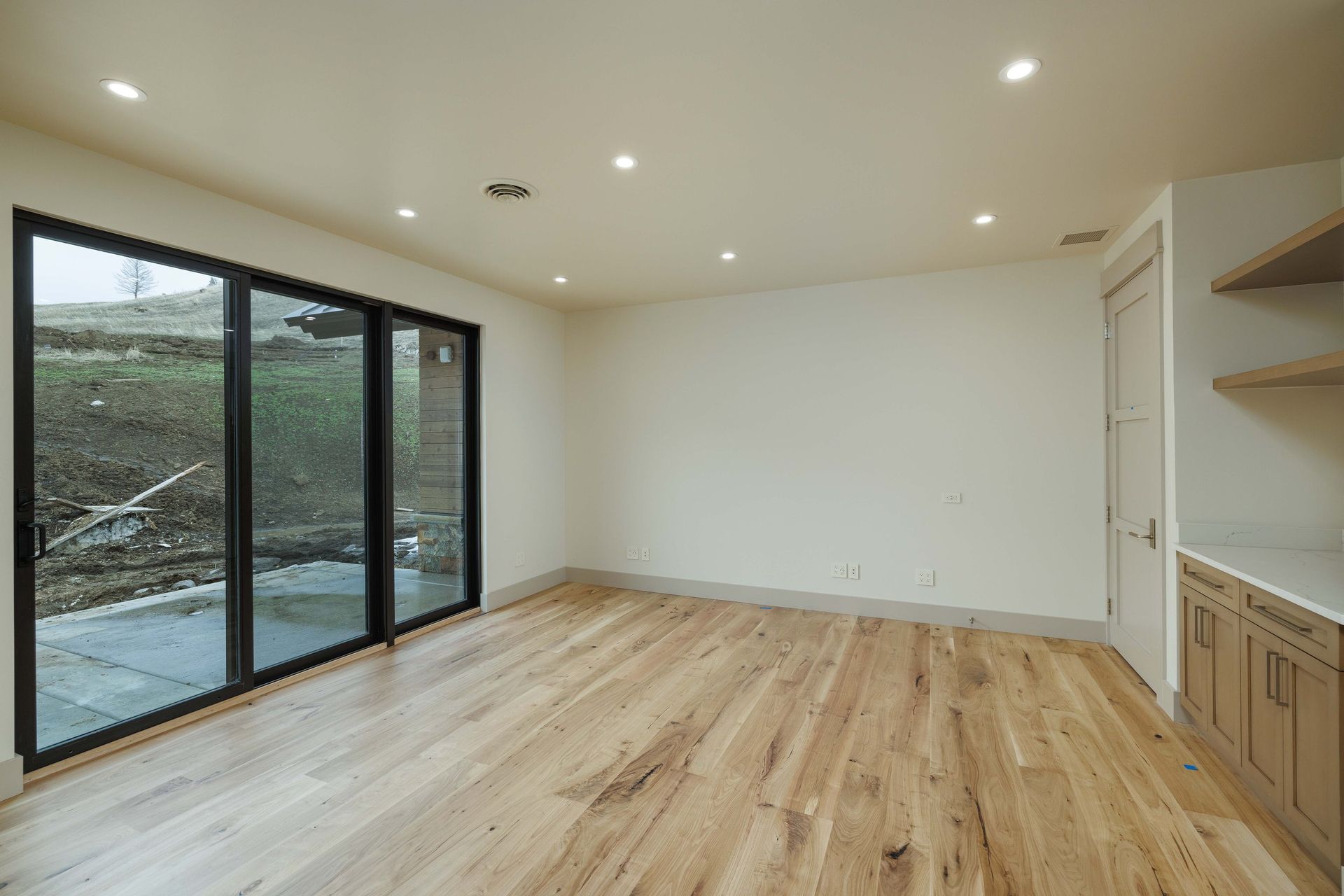 A bright, minimalist room with light wood floors, a large glass sliding door, white walls, and built-in wooden cabinets.