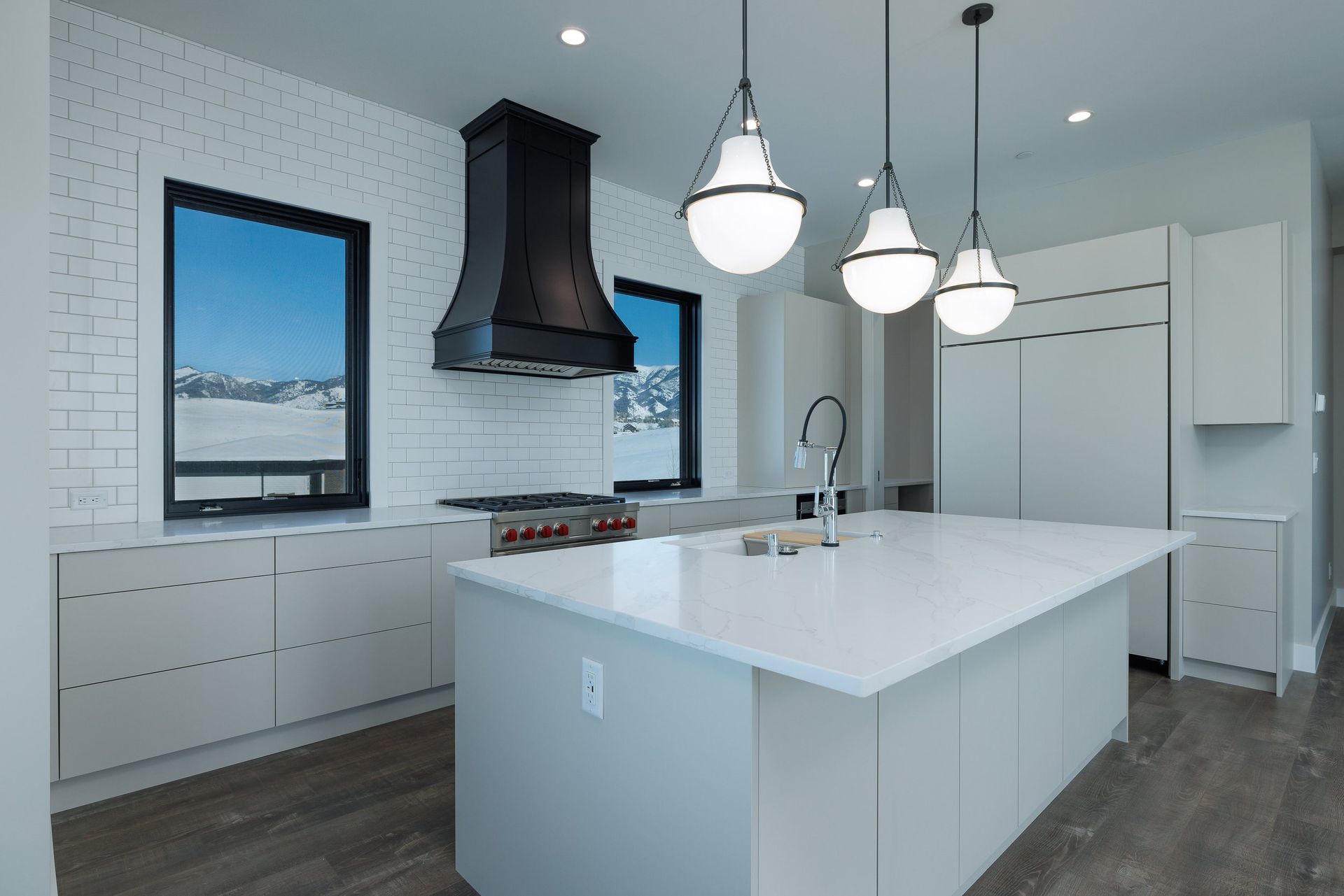 A modern kitchen with light gray cabinets, white countertops, a large black range hood, and three hanging pendant lights.
