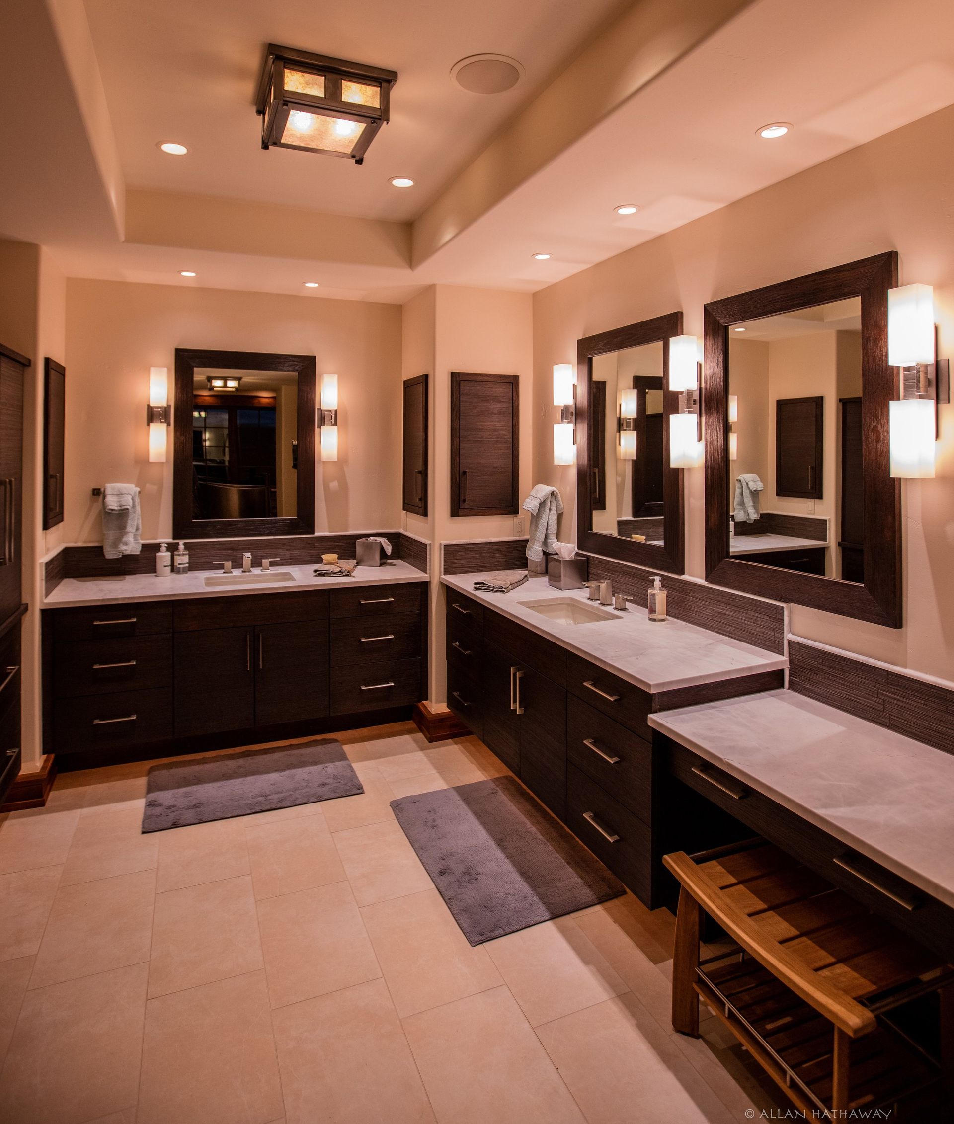 A bathroom with dark wood cabinets, dual sinks, stone countertops, two framed mirrors, and wall sconces on tiled floors.