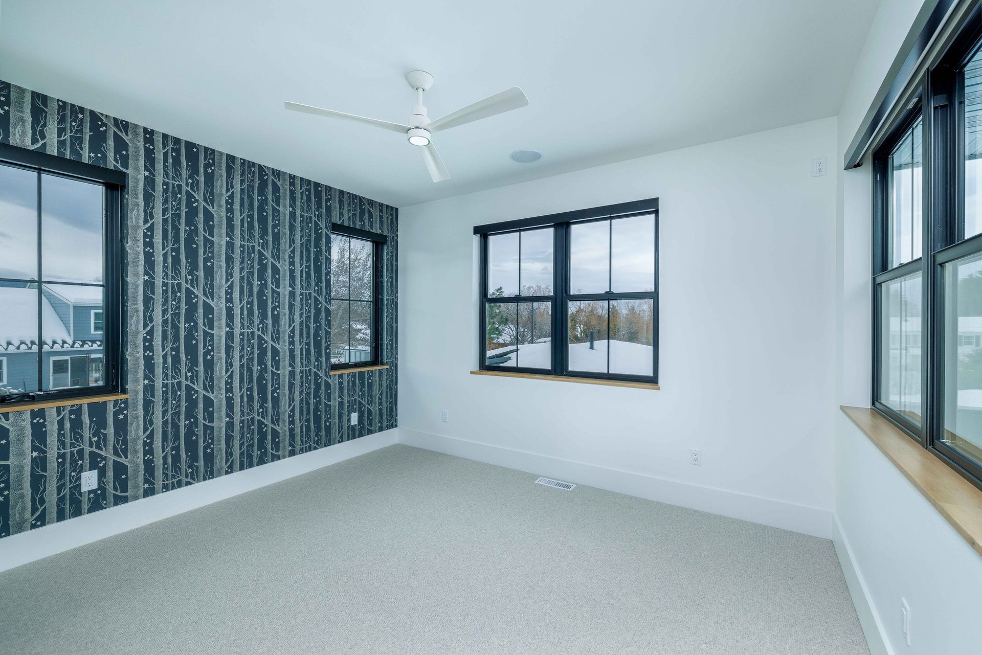 An empty bedroom with a forest-patterned accent wall, light gray carpet, and multiple windows with black frames.