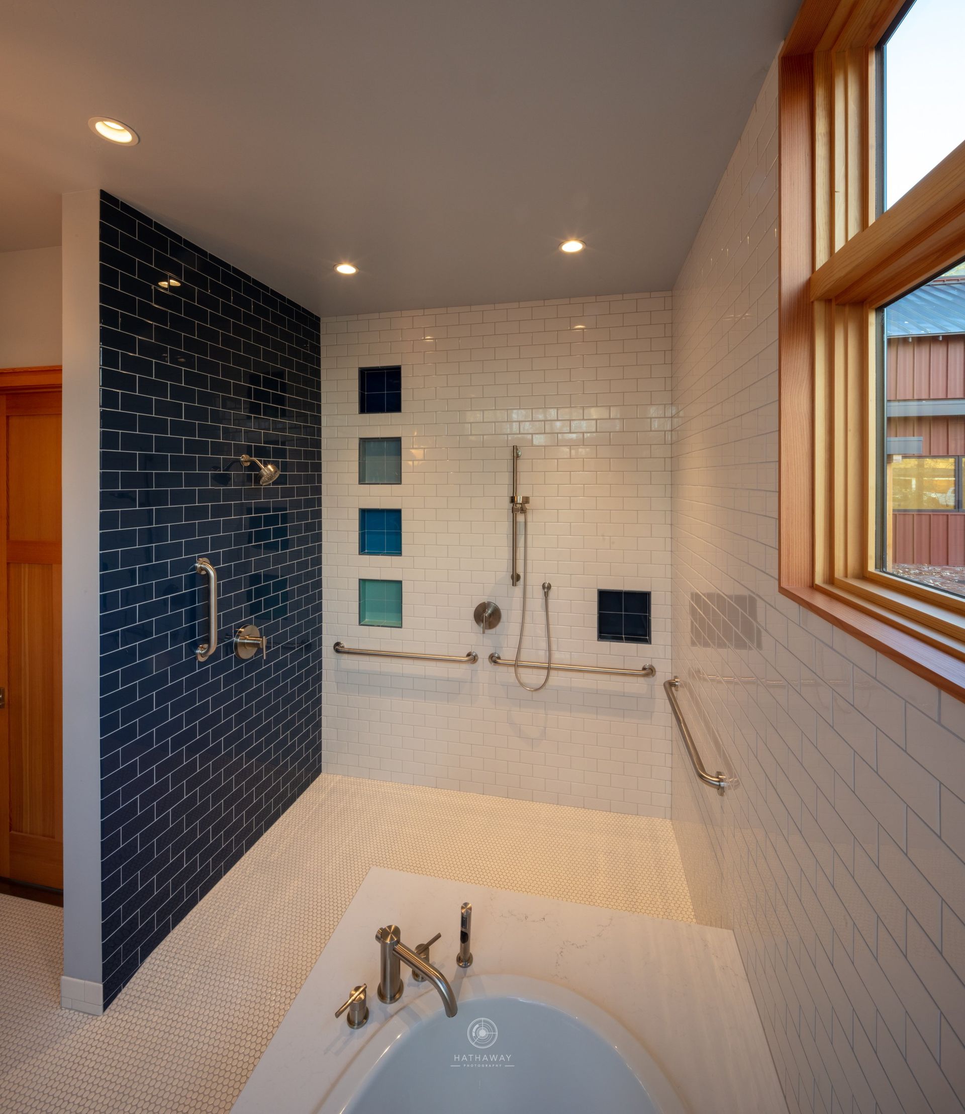 A modern, accessible walk-in shower with blue and white subway tile, grab bars, a handheld sprayer, and a soaking tub.