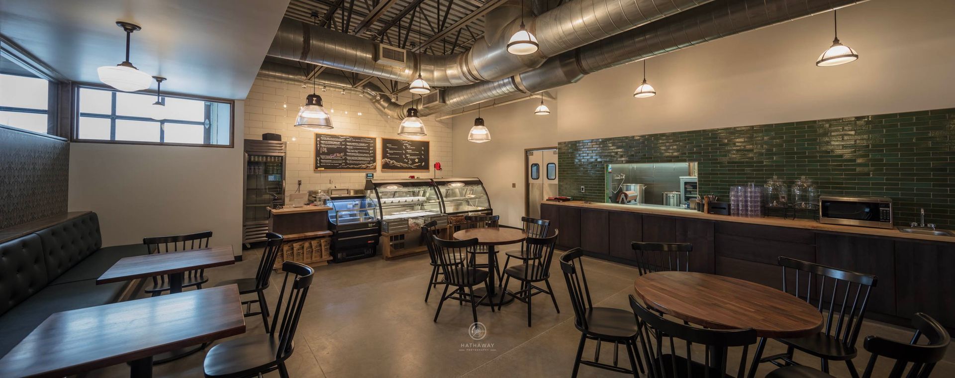 A modern coffee shop interior with tables, chairs, a glass pastry display case, and industrial ceiling ductwork.